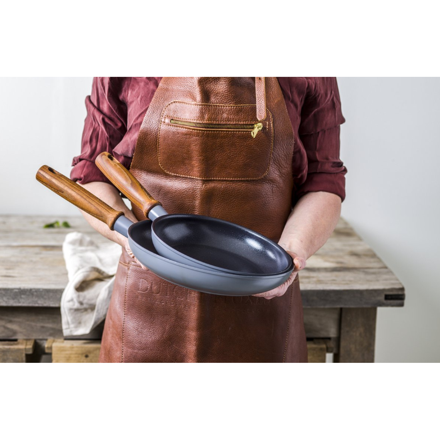 A person wearing a brown apron holds two grey frying pans. Background includes a wooden table and a white cloth.