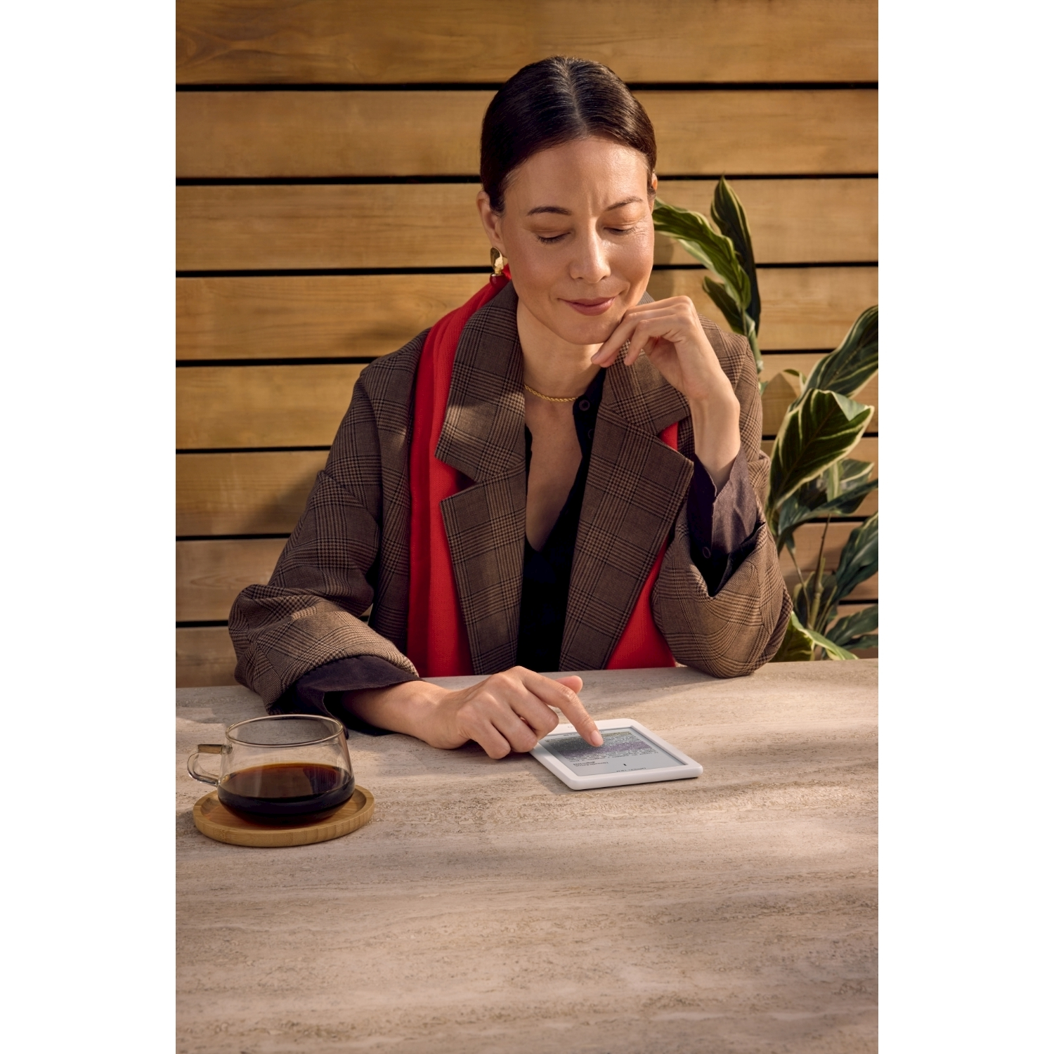 Donna al tavolo, tocca un tablet. Tazza di caffè a sinistra. Parete in legno sullo sfondo. Piante a destra.