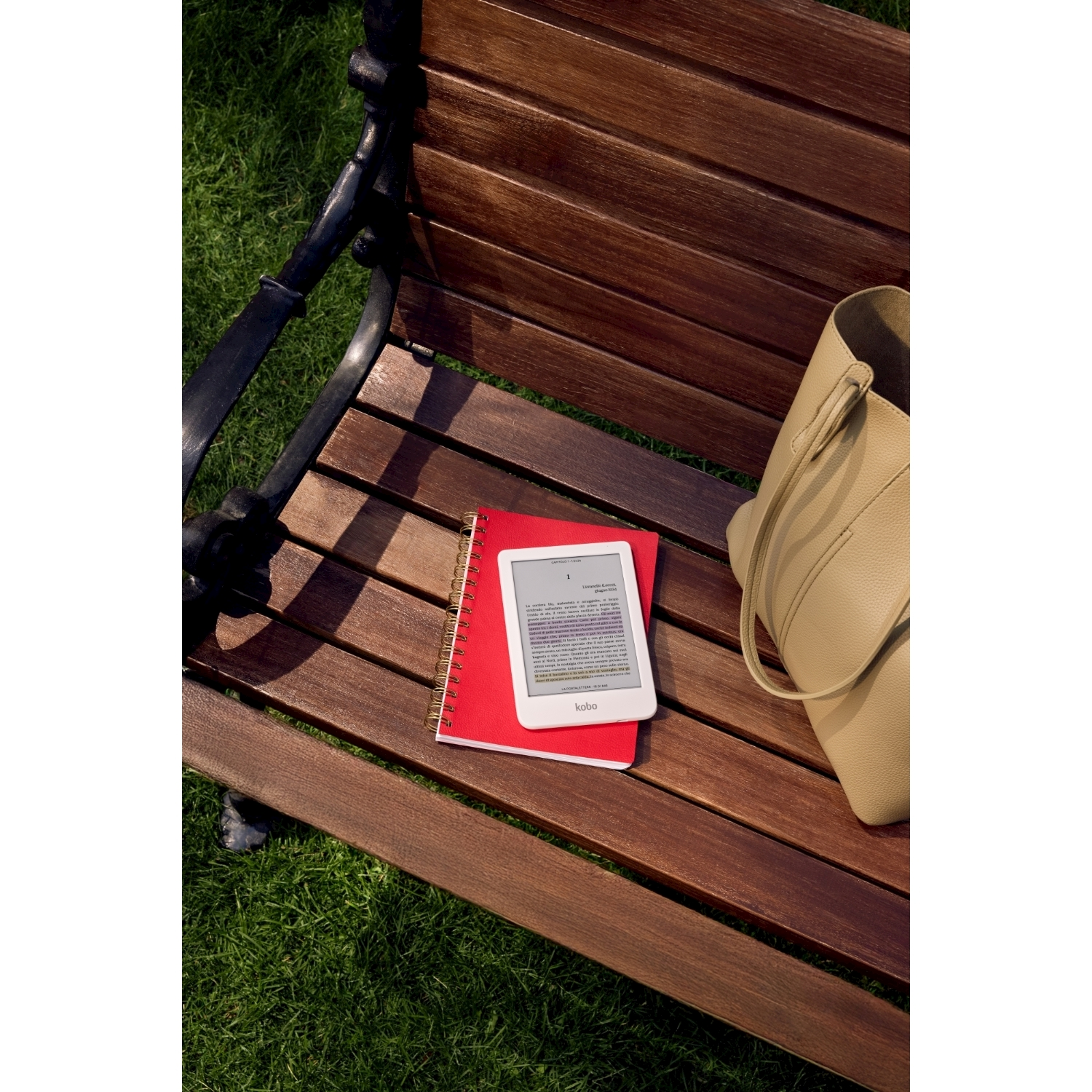 Una panchina di legno con un taccuino rosso, un e-reader bianco e una borsa beige. Erba verde sullo sfondo.