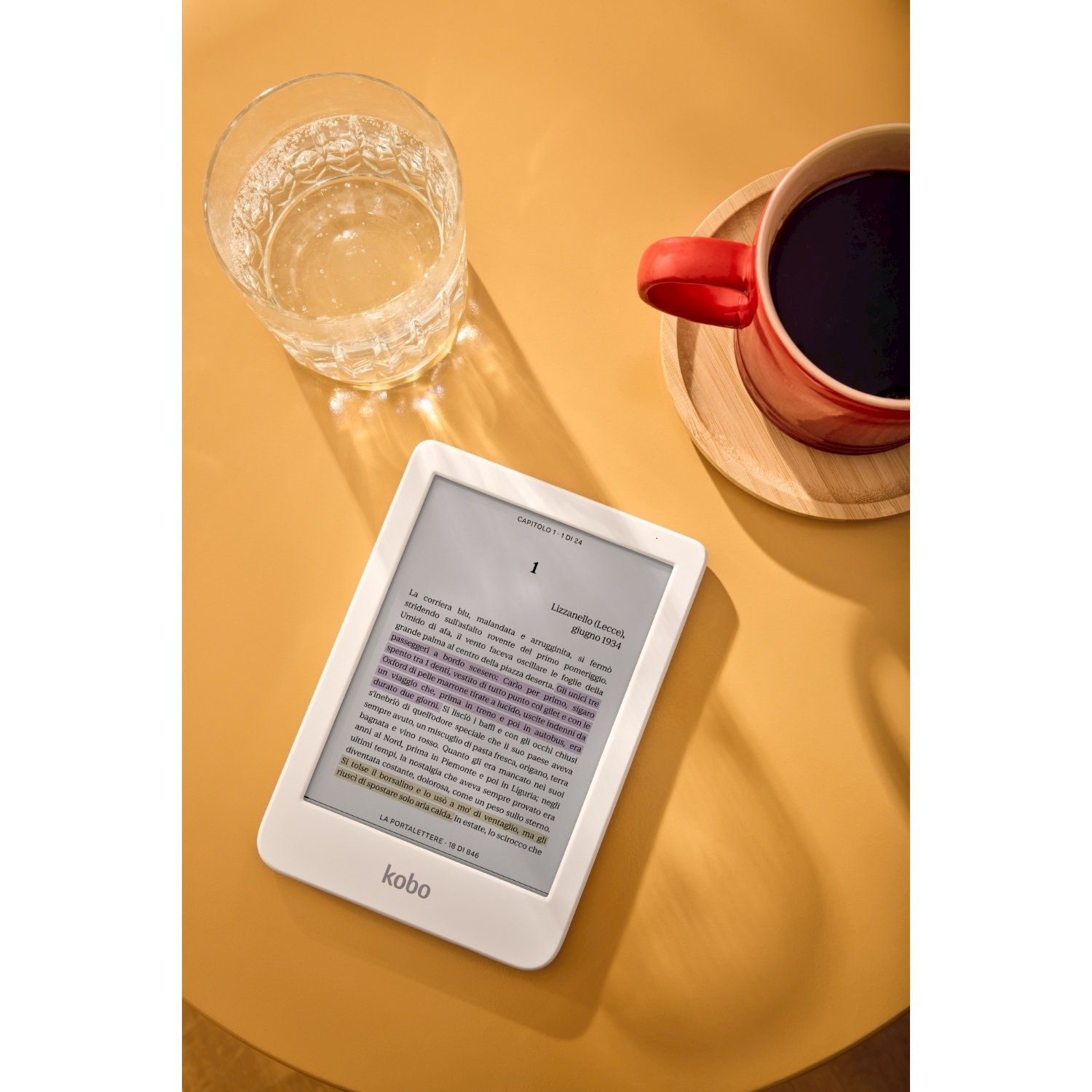 Un e-reader bianco, un bicchiere d'acqua e una tazza di caffè rossa su un tavolo giallo.