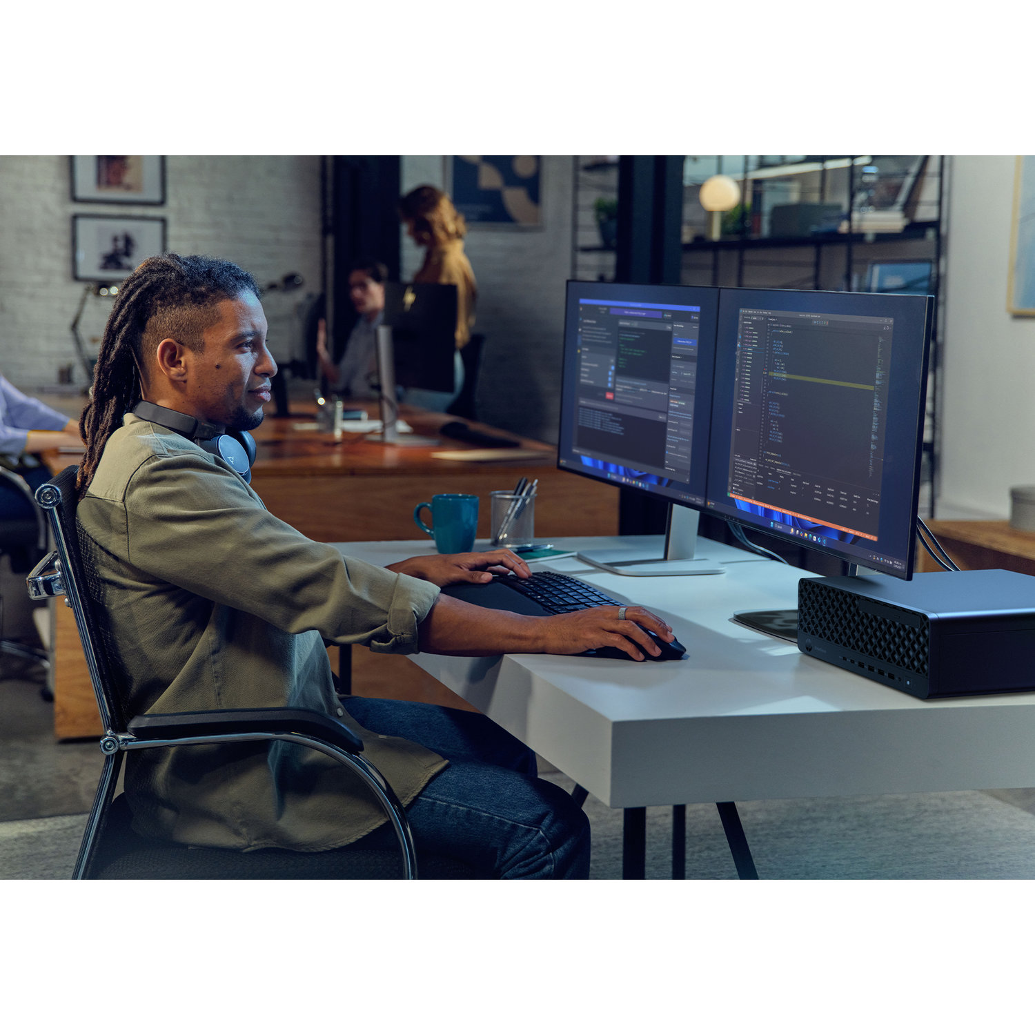 A man with dreadlocks works at a desk with two monitors. He types on a keyboard. Other people work at desks in the background.