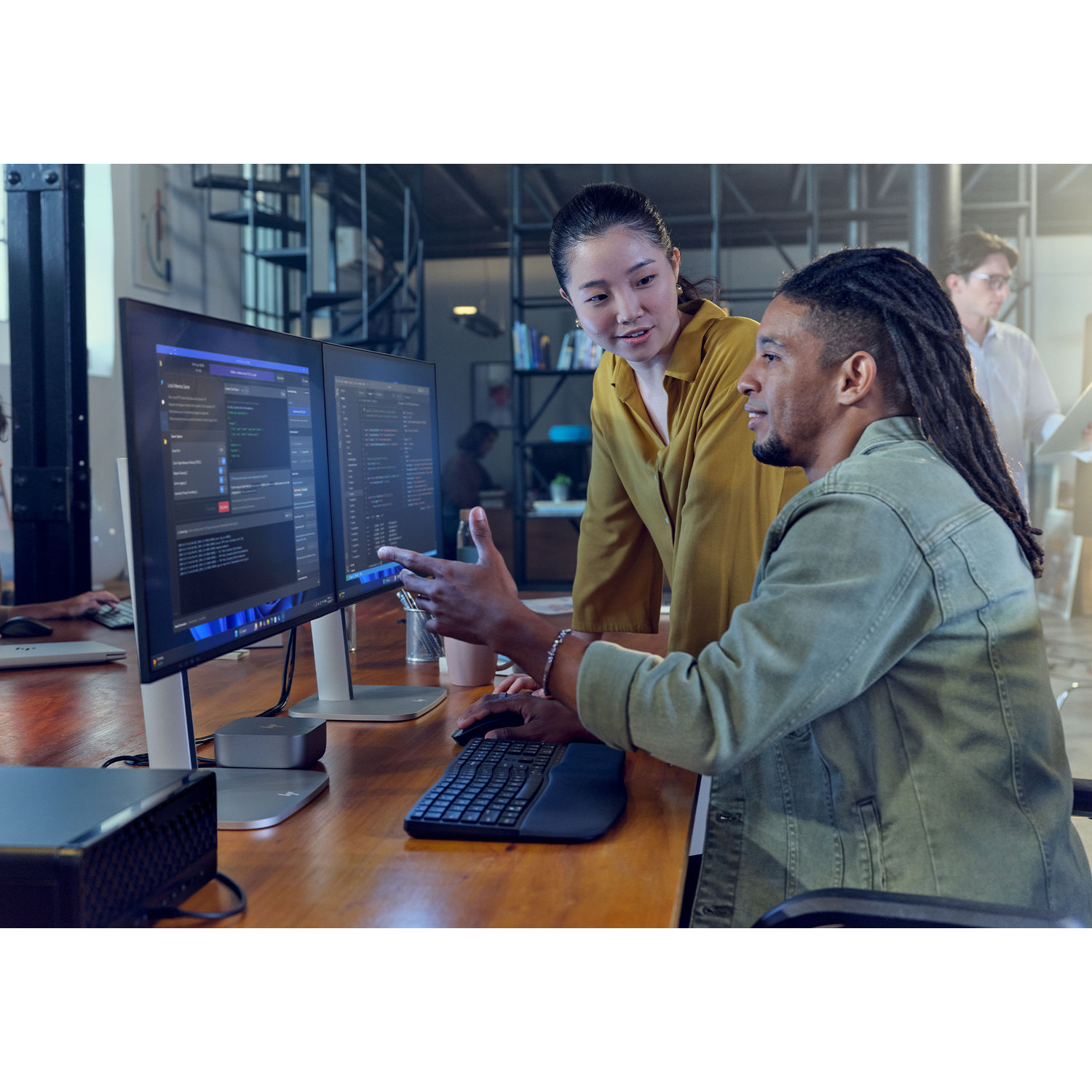 Two people looking at computer screens. One points, discussing code. A third person stands in the background.