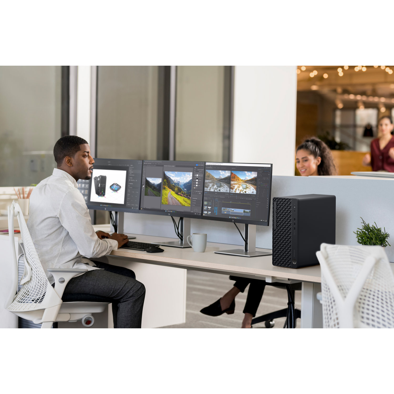Een man werkt aan een bureau met drie monitoren. Er zijn nog twee vrouwen op de achtergrond. Er staat ook een computer op het bureau.