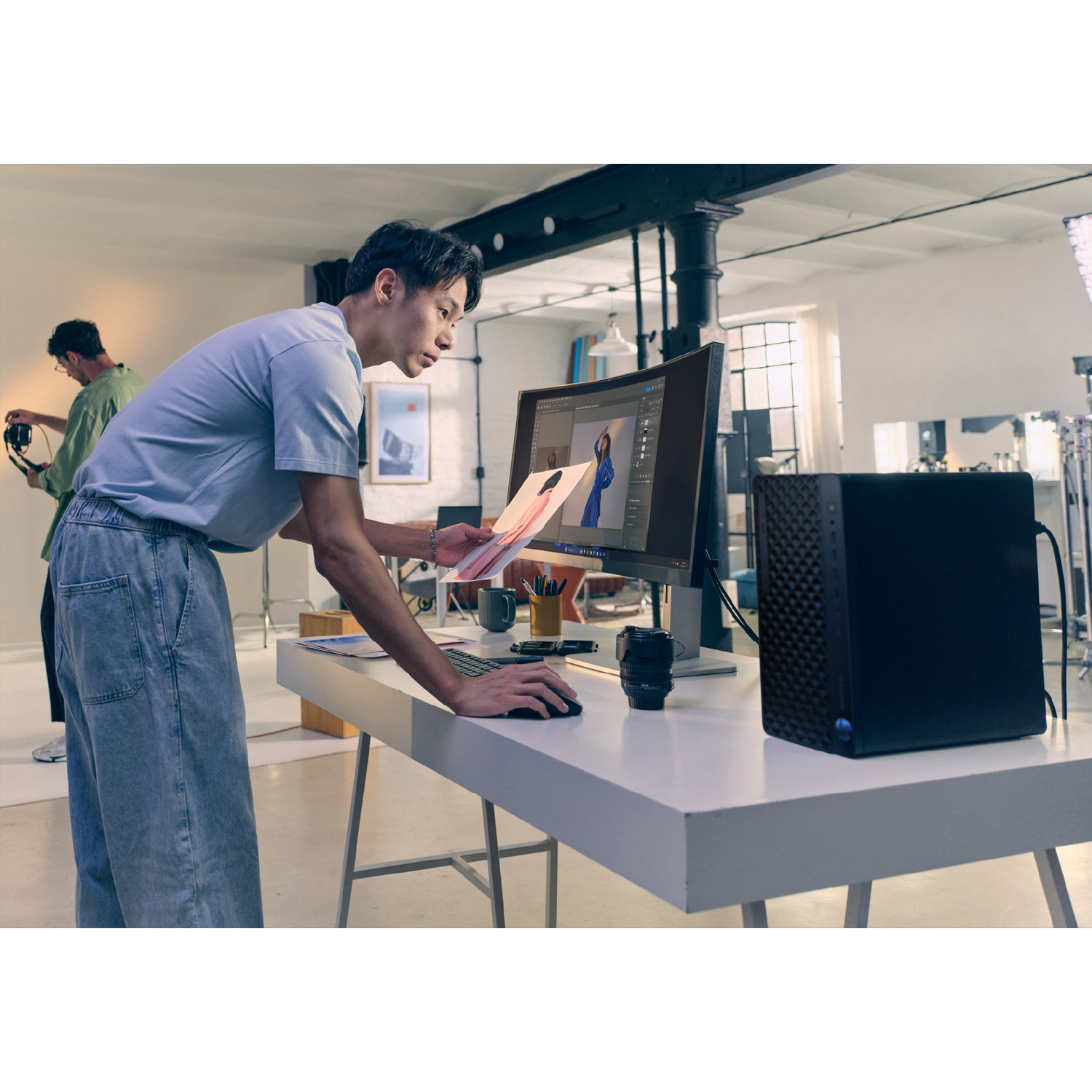 Een man bekijkt een fotoafdruk bij een computer, terwijl een ander persoon een camera bedient op de achtergrond. Er staat een zwarte computertoren op het bureau.