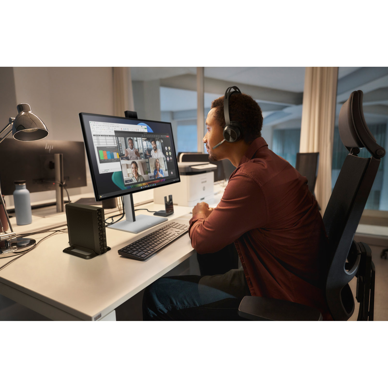 Een man met een headset neemt deel aan een videoconferentie aan zijn bureau. Een computerscherm toont de video-oproep. Toetsenbord en kleine pc op het bureau.