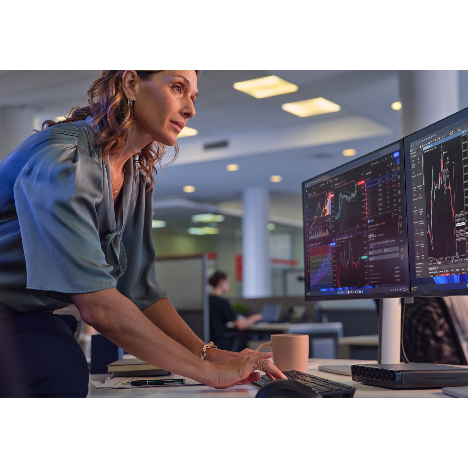 Een vrouw in een blauw overhemd bekijkt twee computerschermen met aandelenkoersen. Een mok, een toetsenbord en een computer staan op het bureau.