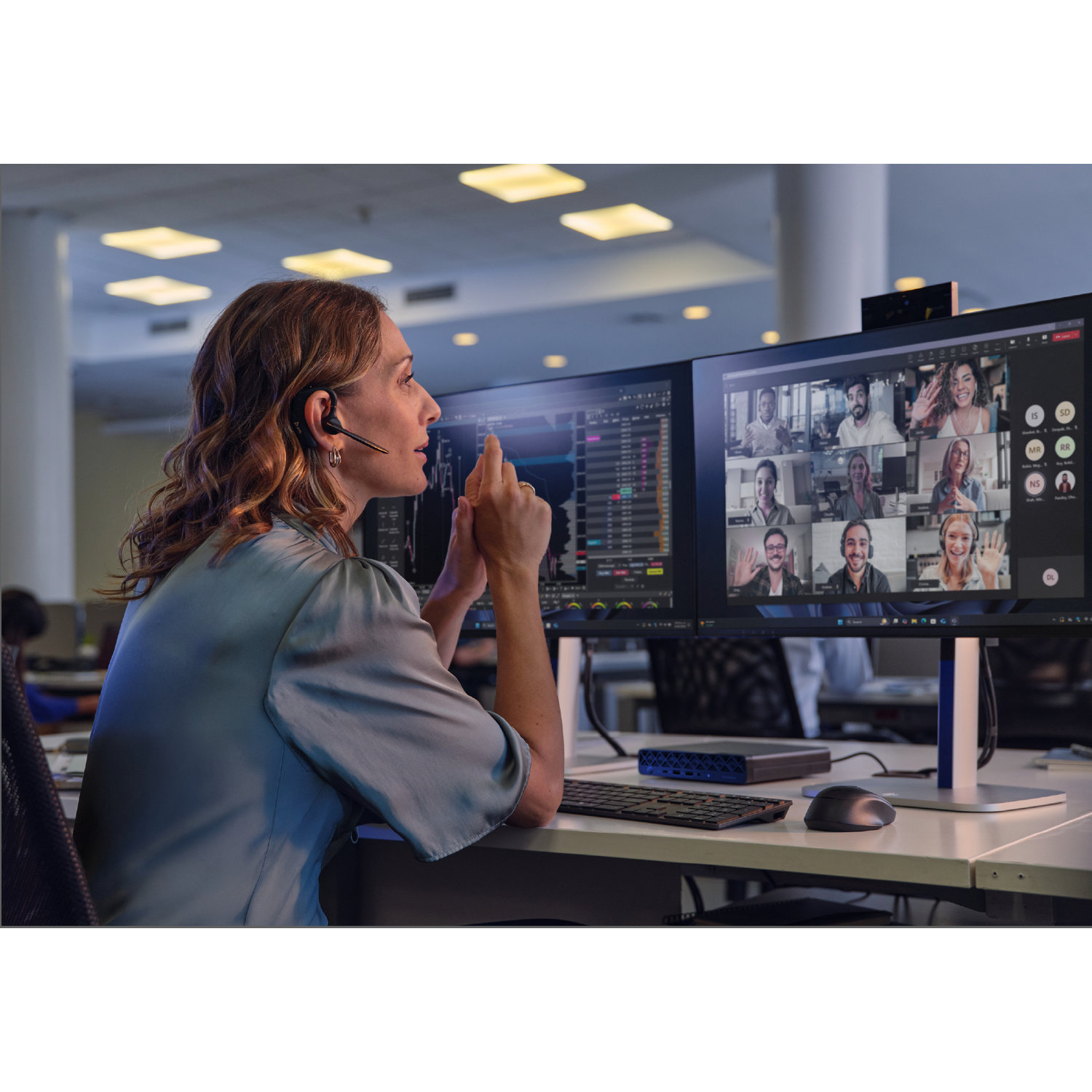 Vrouw met headset in een videogesprek, bekijkt een scherm met meerdere mensen. Toetsenbord, muis en monitor op het bureau.