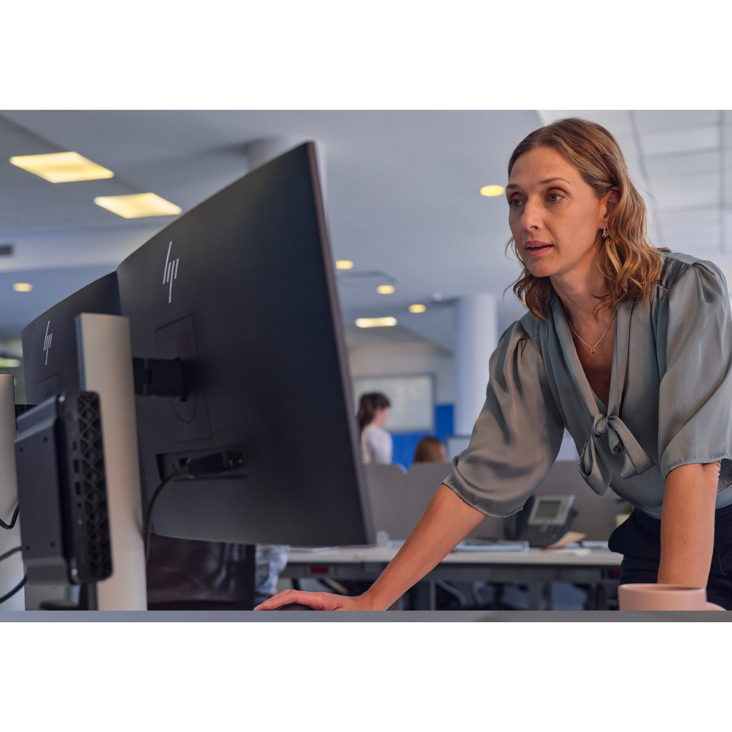 Een vrouw in een teal blouse kijkt naar een computerscherm. Ze leunt op een bureau in een kantooromgeving.