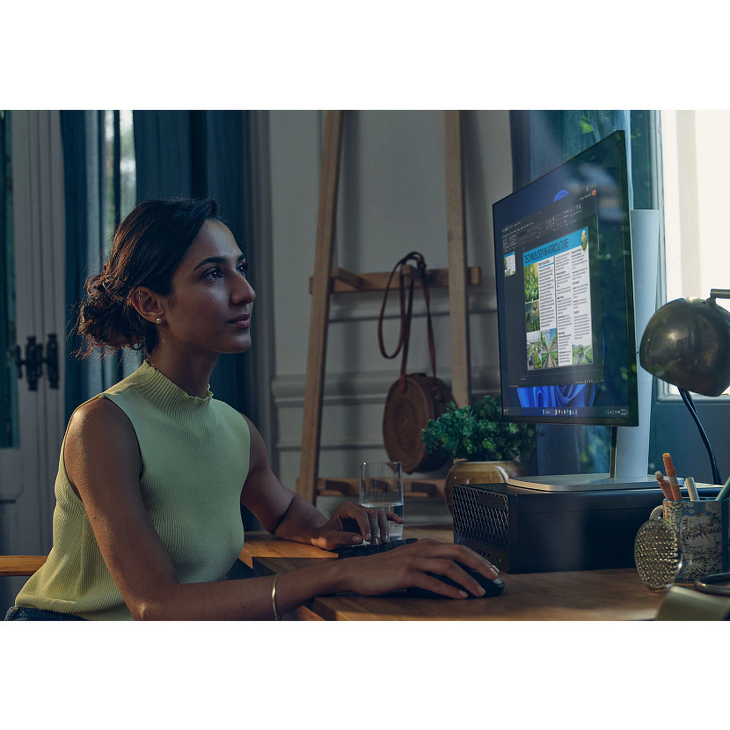 A woman works on a computer. She wears a light green top. A monitor displays a document. A glass of water is on the desk.