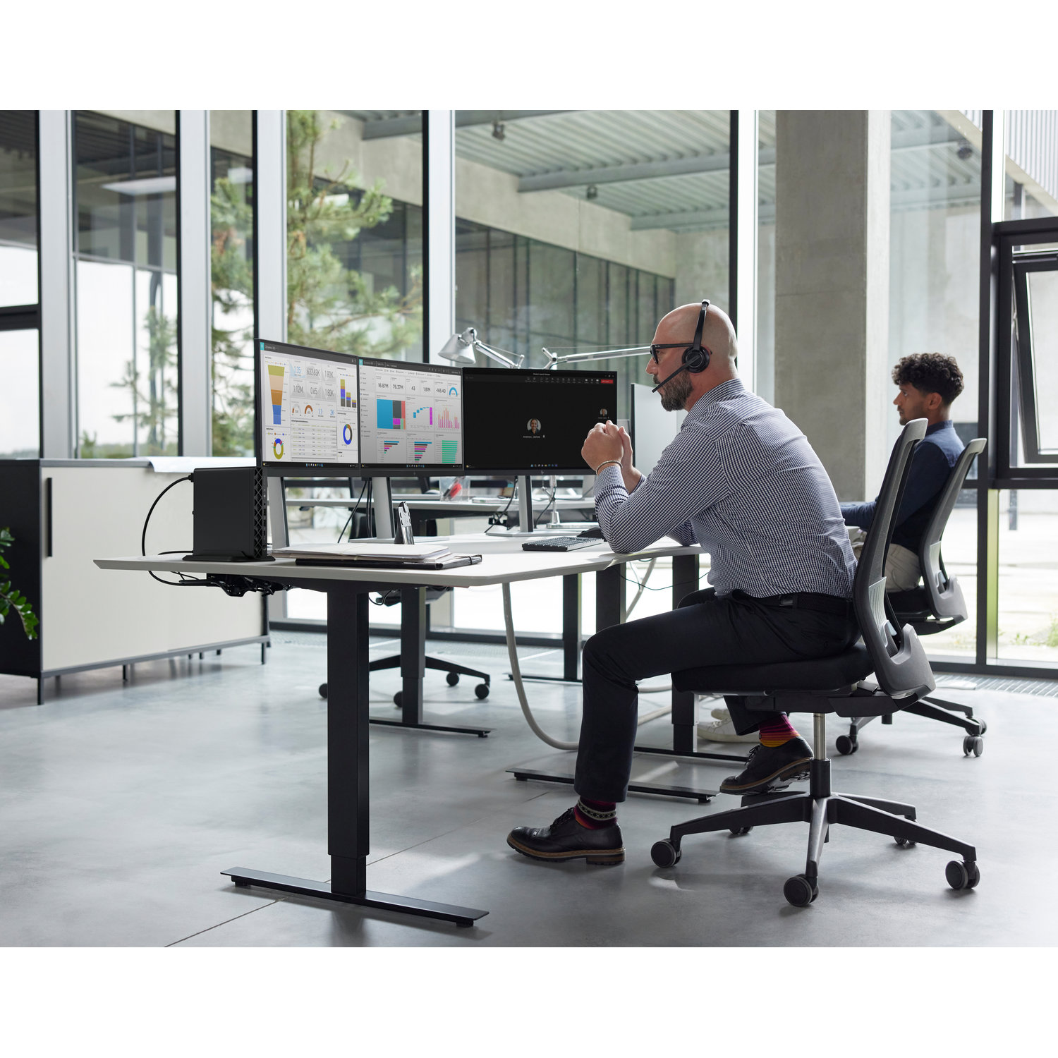 Two men in an office, one wearing a headset, looking at computer screens displaying charts. Another man sits at a desk.
