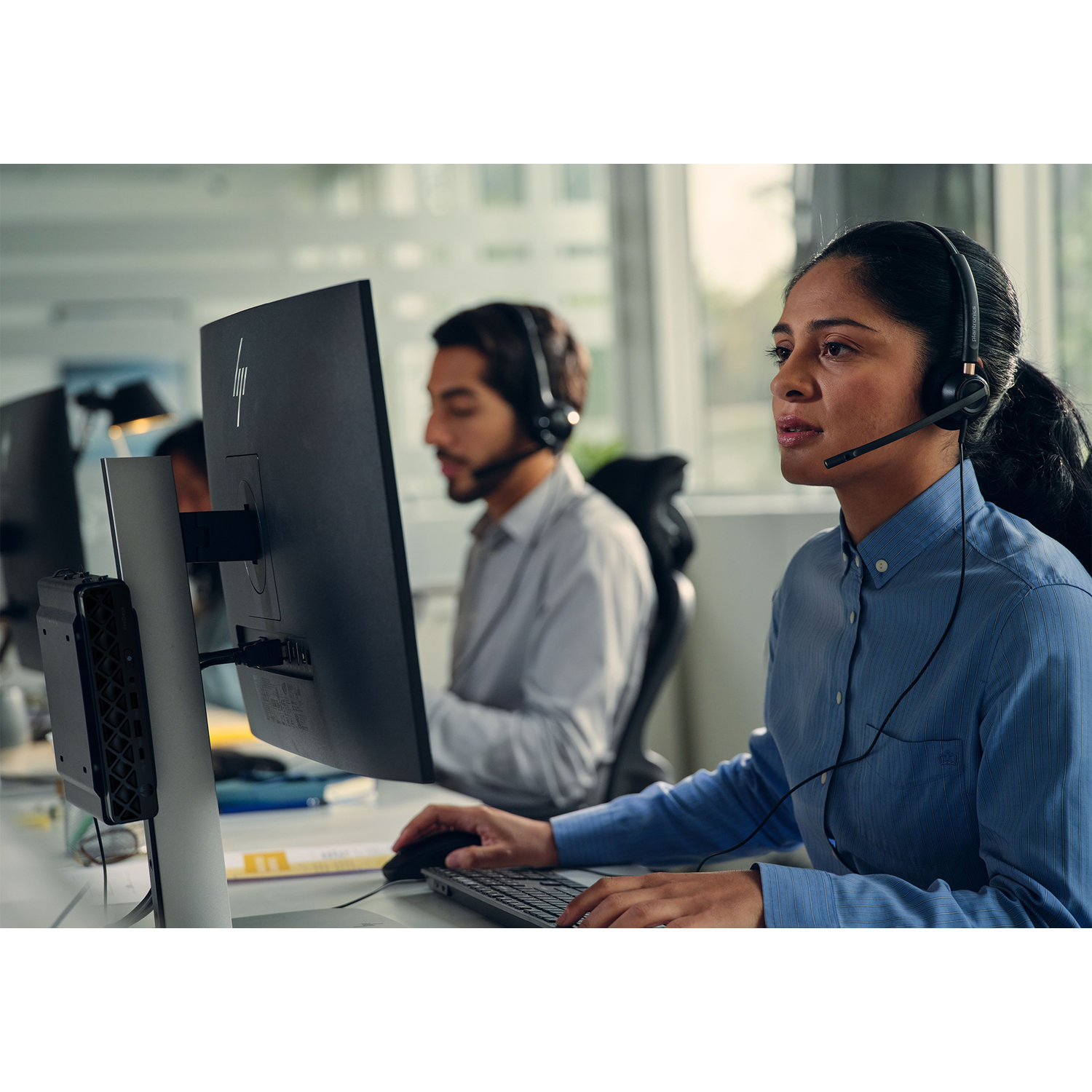 A woman in a blue shirt wears a headset, working on a computer. A man in the background also wears a headset.