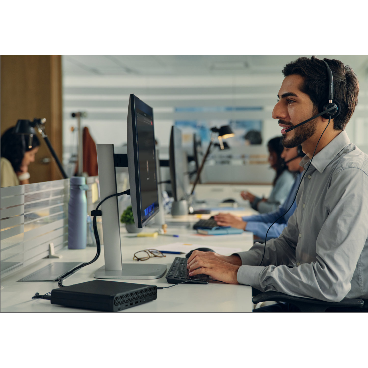 A man wearing a headset is working on a computer. Others are in the background. A black device sits on the desk.