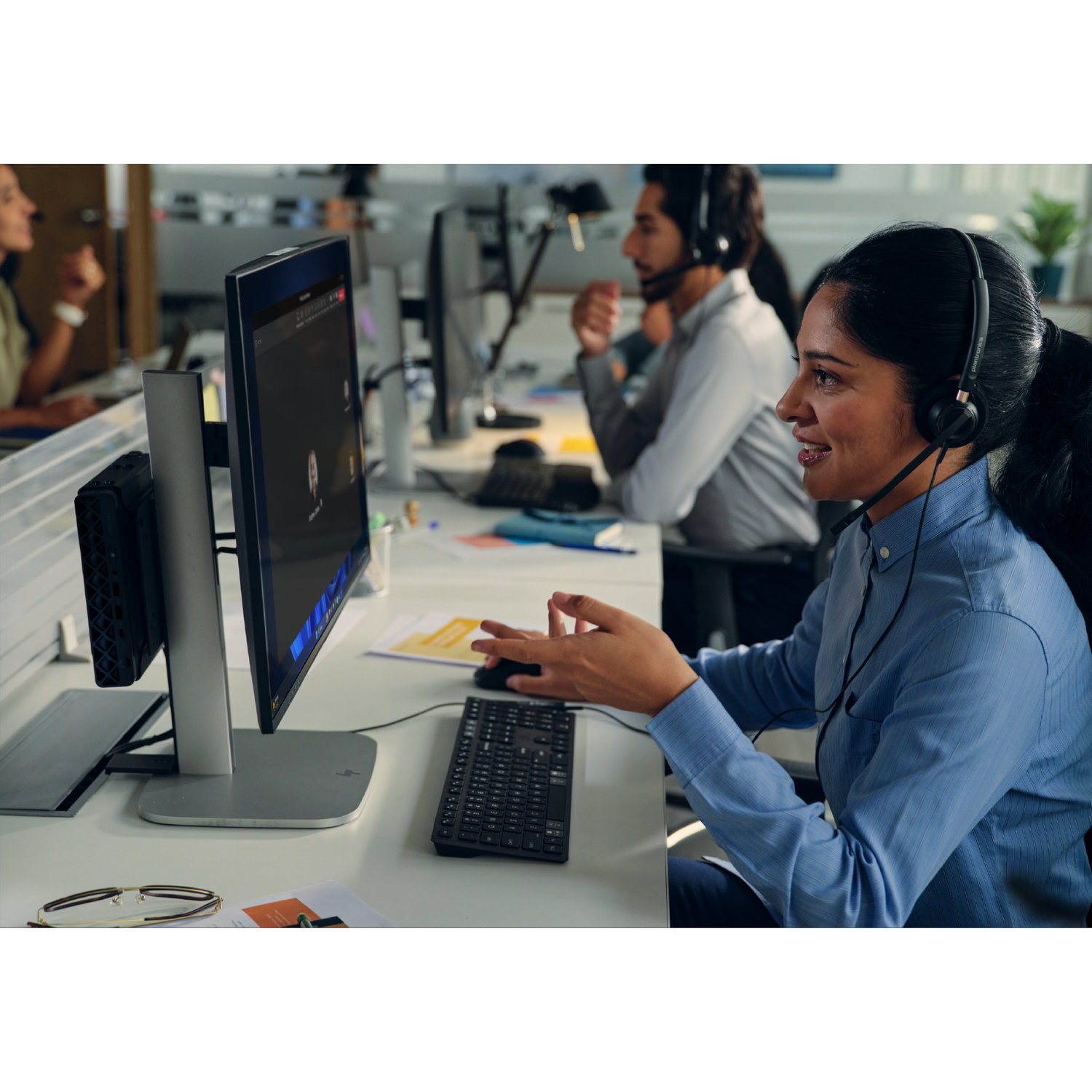 A woman wearing a headset gestures at a computer screen. Others in the background wear headsets at their desks.
