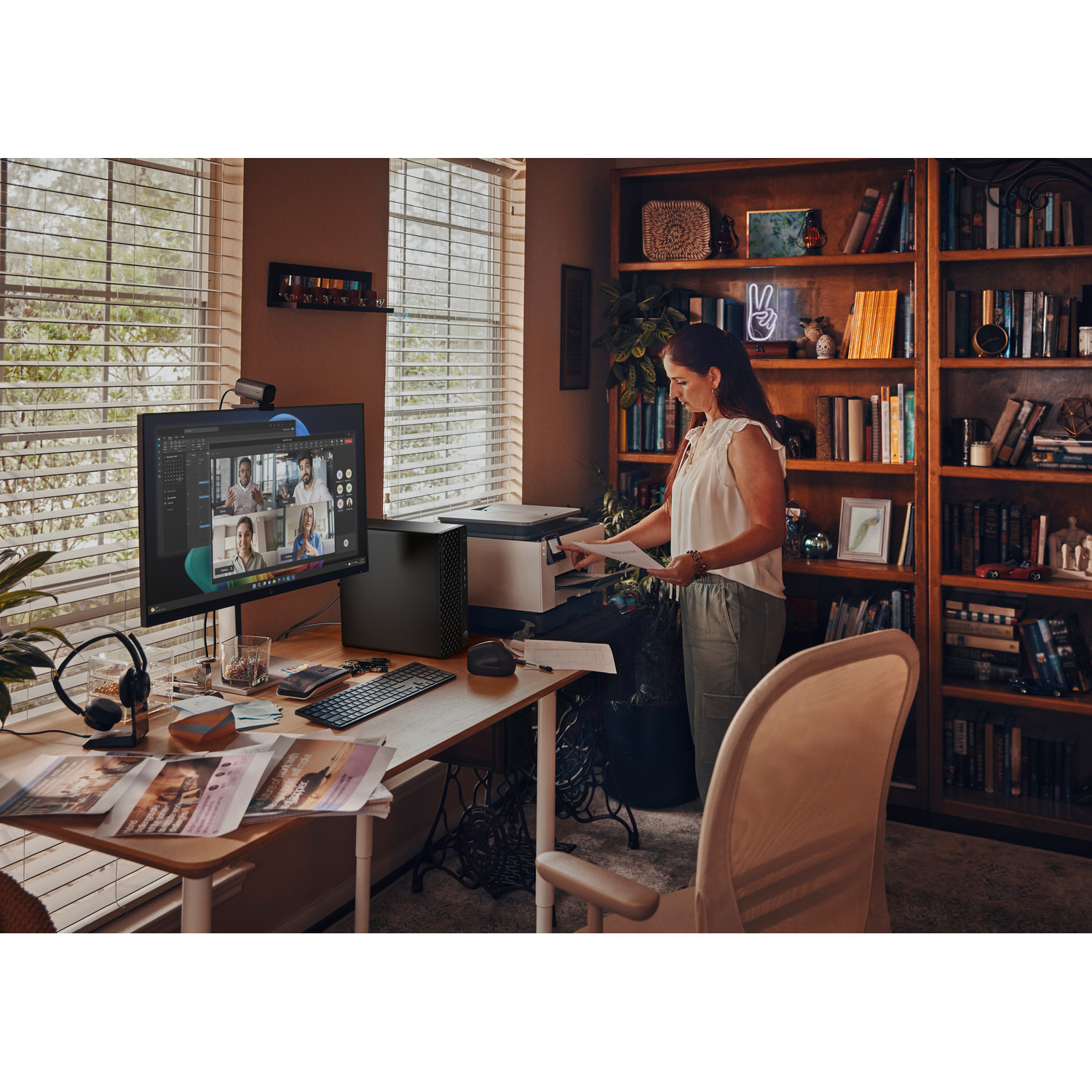 A woman stands near a desk in a home office. A monitor displays a video call. Bookshelves and a window are in the background.