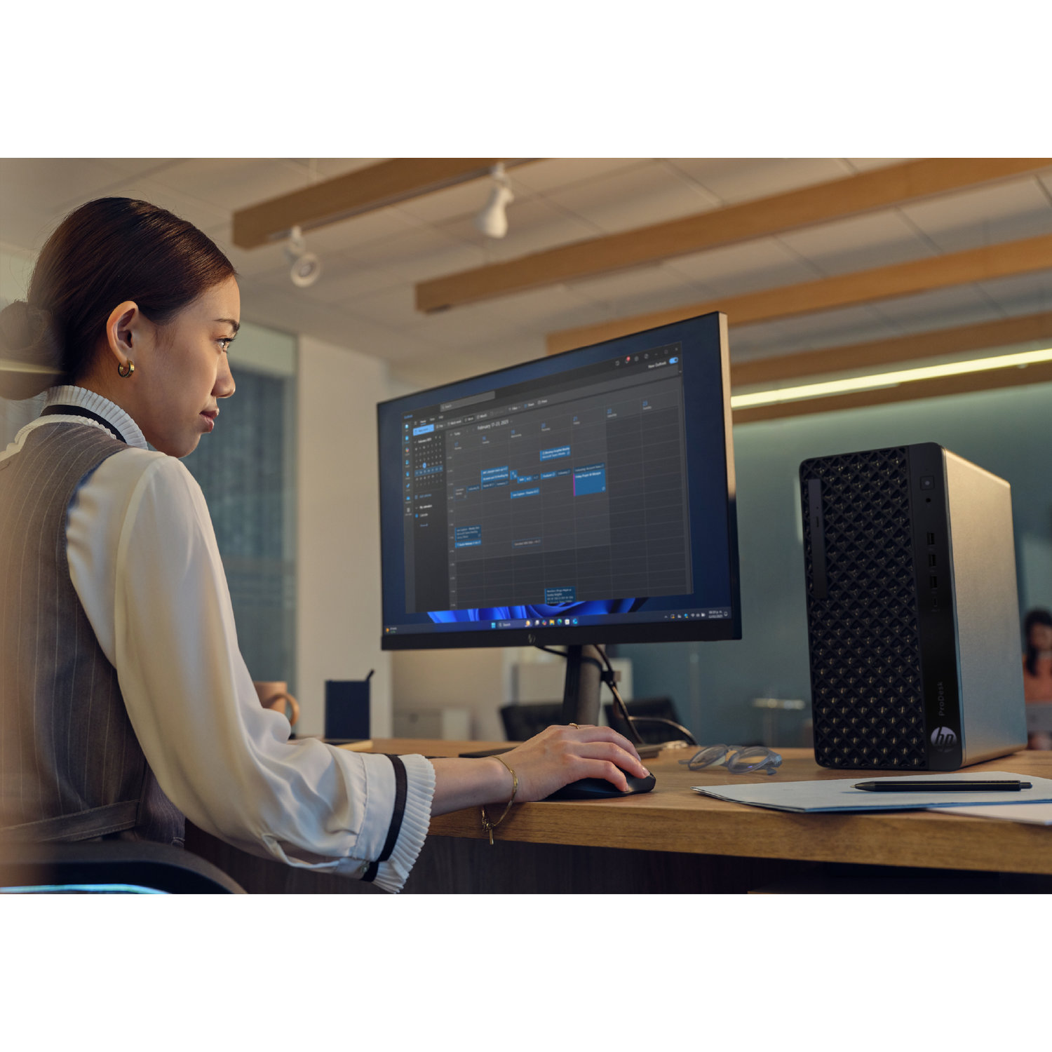 A woman works at a desk, using a computer showing a calendar. A computer tower and glasses are beside the monitor.