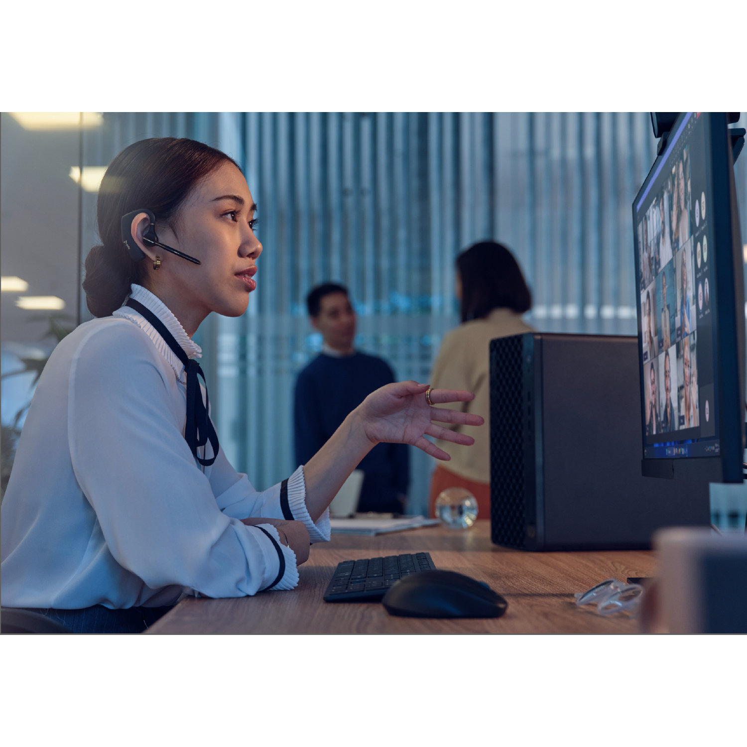 Woman in headset gestures towards a computer screen with a video call. Two people are in the background.