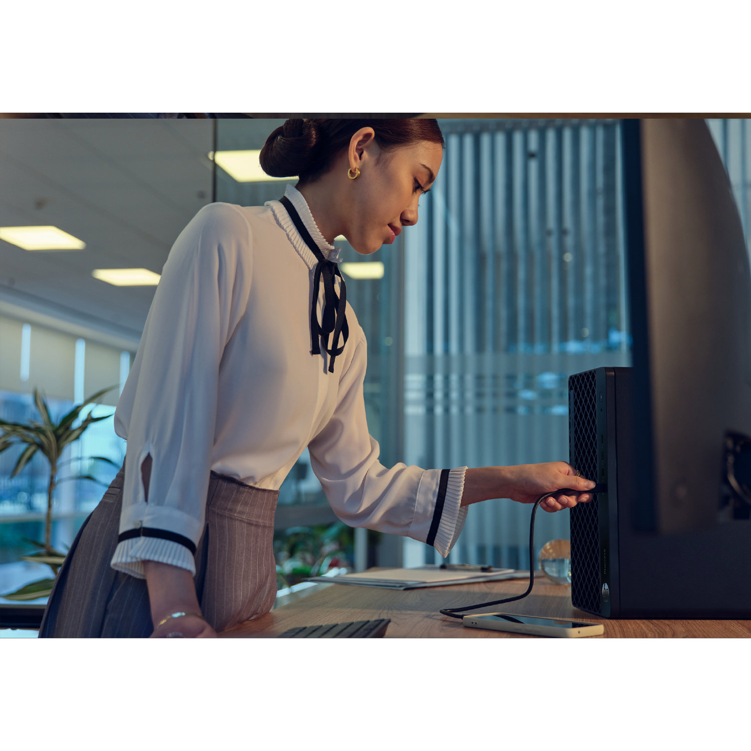 A woman connects a cable to a black computer tower in an office. She wears a white blouse and gray skirt.
