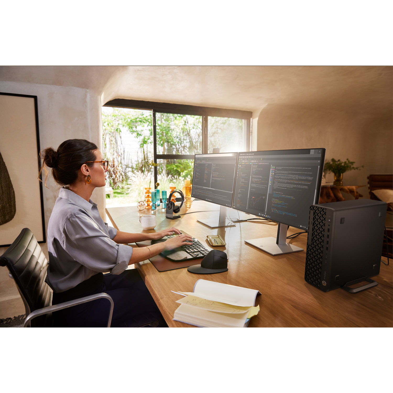 A woman works on a computer with two monitors. A black computer tower sits on the desk. A window with greenery is in the background.