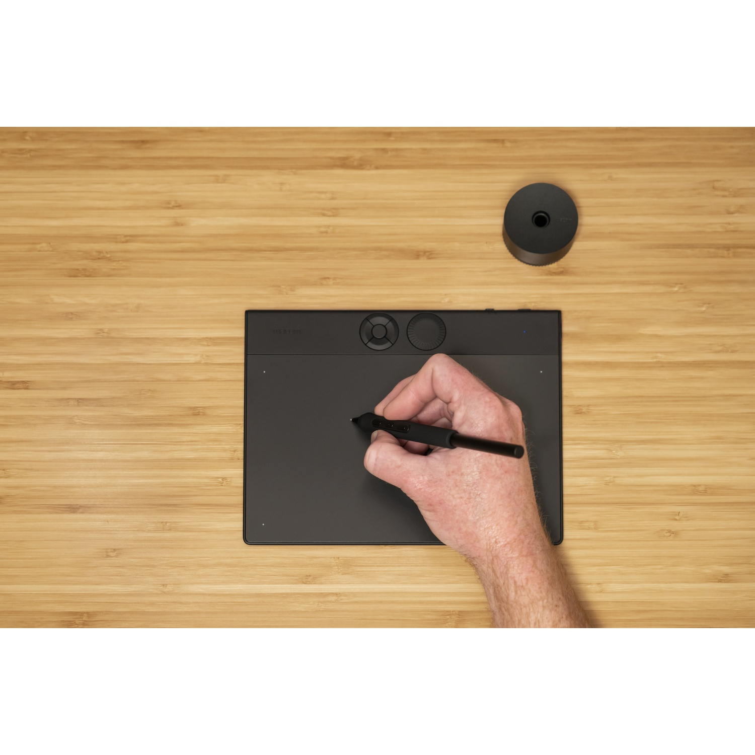 Una mano che tiene una penna su un tablet nero su una superficie di legno. Un cilindro nero è in alto a destra.