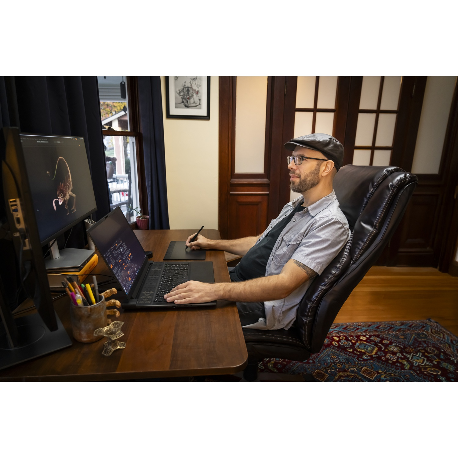 Uomo con occhiali e cappello lavora su laptop, tavoletta grafica e monitor. È seduto a una scrivania di legno.