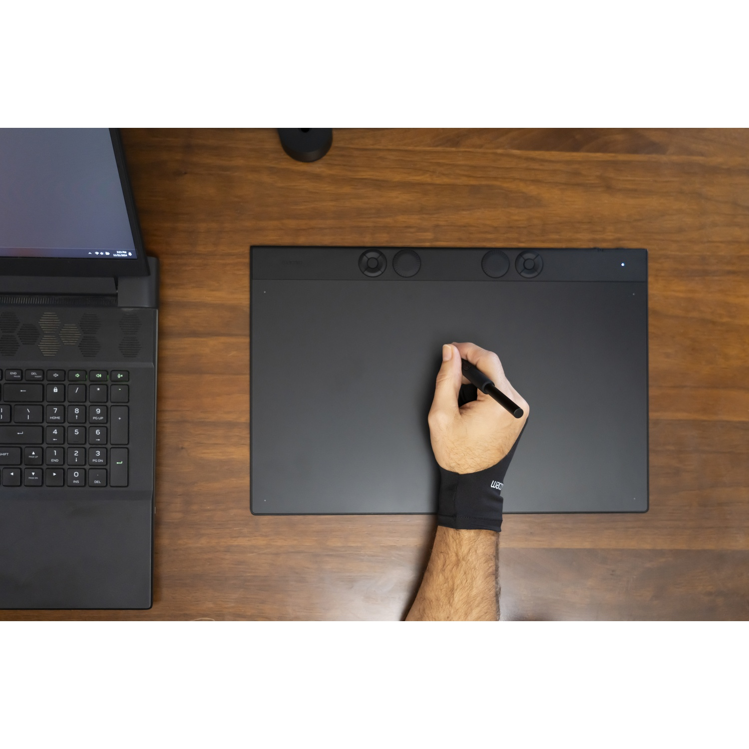 Una persona disegna su un tablet con una penna, indossando un guanto nero. Un laptop è a sinistra, tutto su un tavolo di legno.