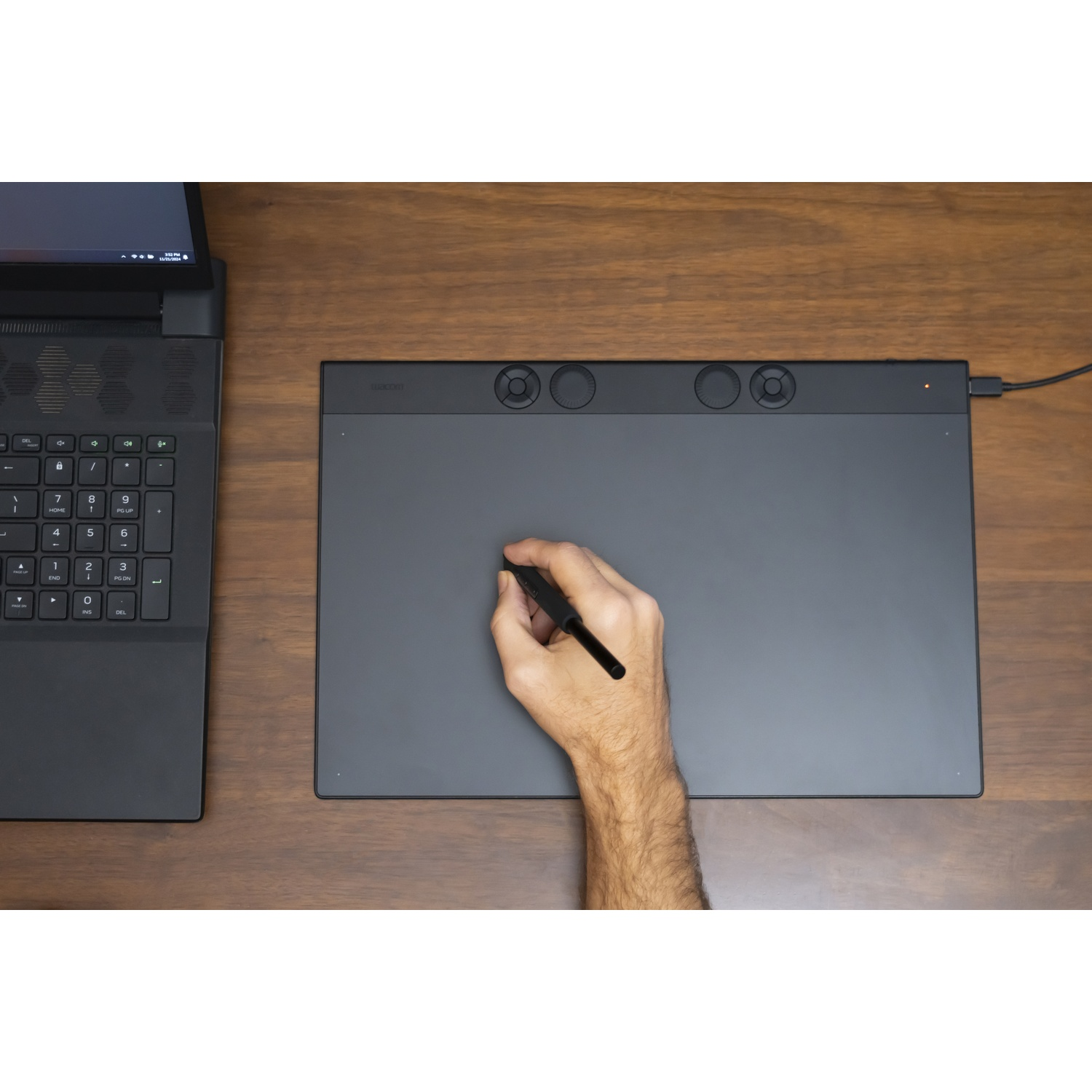 Una mano tiene una penna su una tavoletta grafica. Un laptop è a sinistra. La tavoletta è su una superficie di legno.