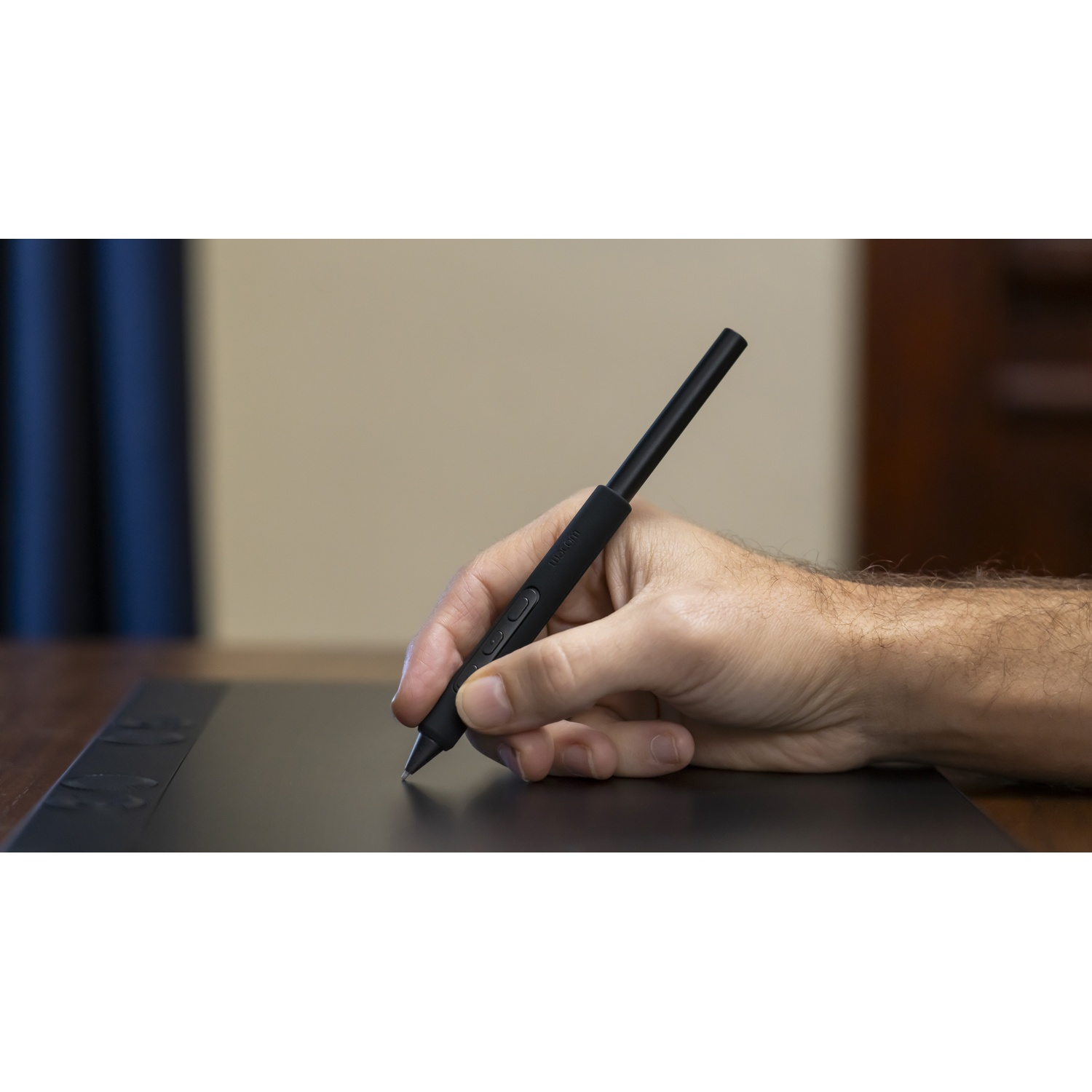 Una mano tiene uno stilo nero, che scrive su un tablet nero. Lo sfondo comprende un tavolo di legno e un muro beige.