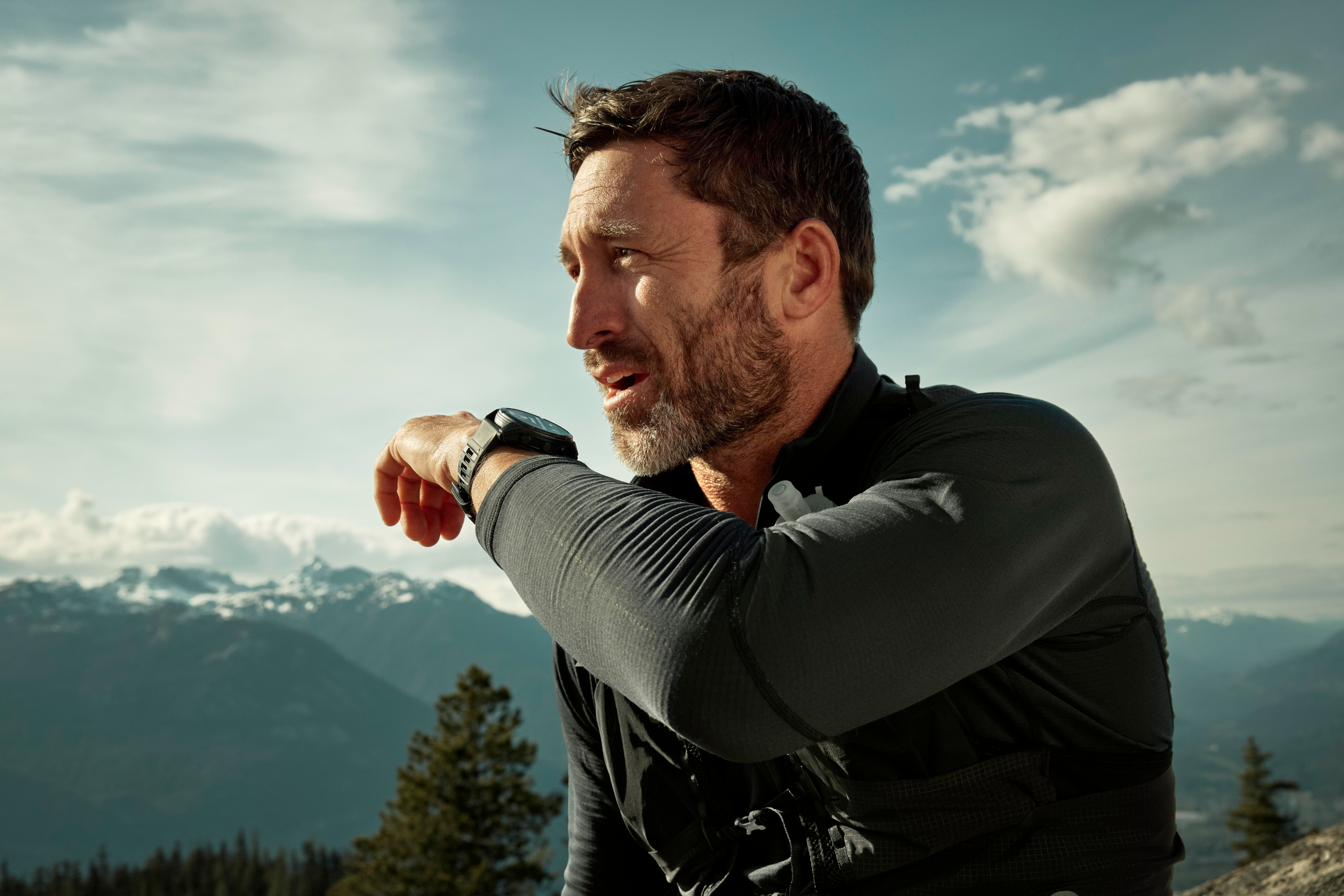 Ein Mann mit Bart und Smartwatch. Er blickt weg, Berge und Himmel im Hintergrund.