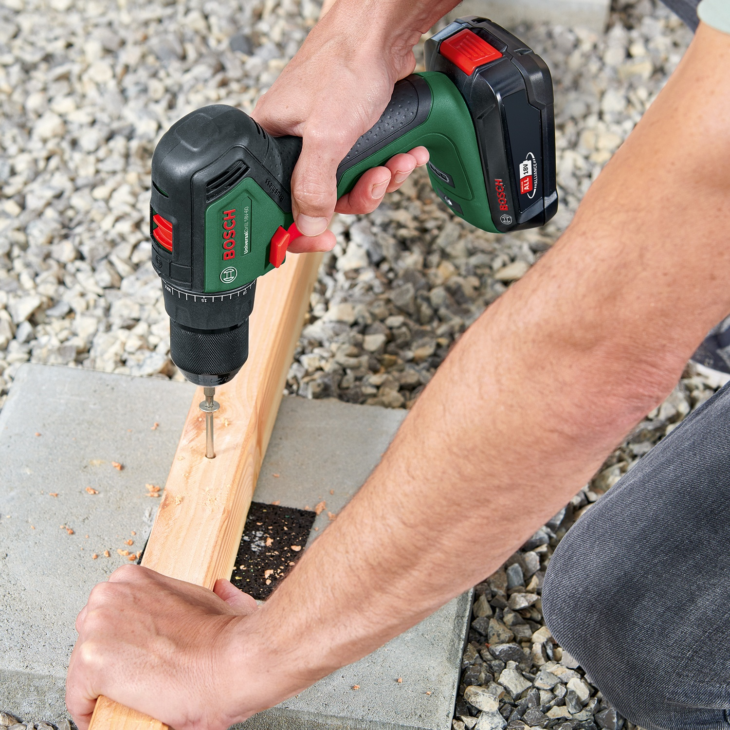 Una persona avvita una vite in una tavola di legno con un trapano avvitatore a batteria Bosch. Ghiaia grigia e lastre di cemento sullo sfondo.
