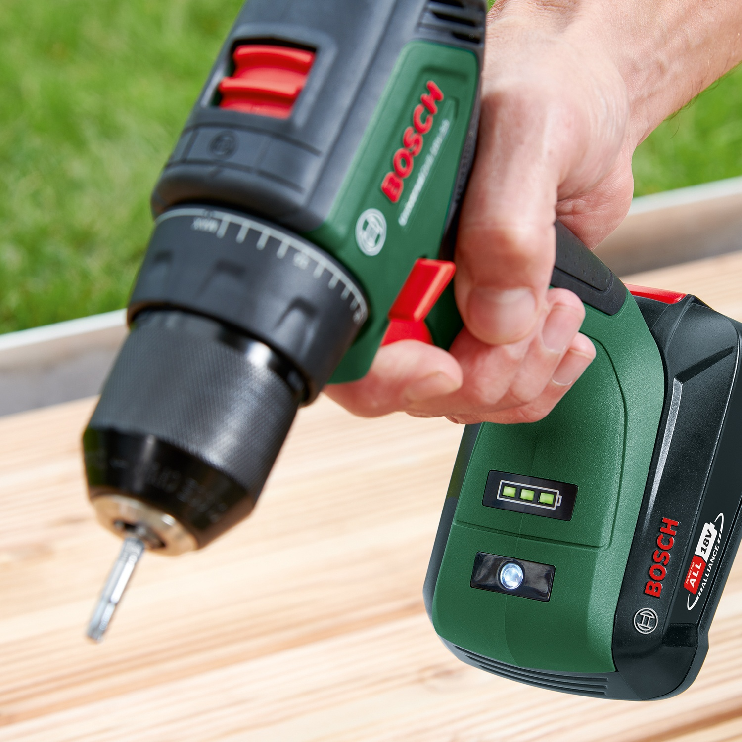 Una mano tiene un trapano Bosch verde e nero. Il trapano è puntato verso una superficie di legno. L'erba verde è sullo sfondo.