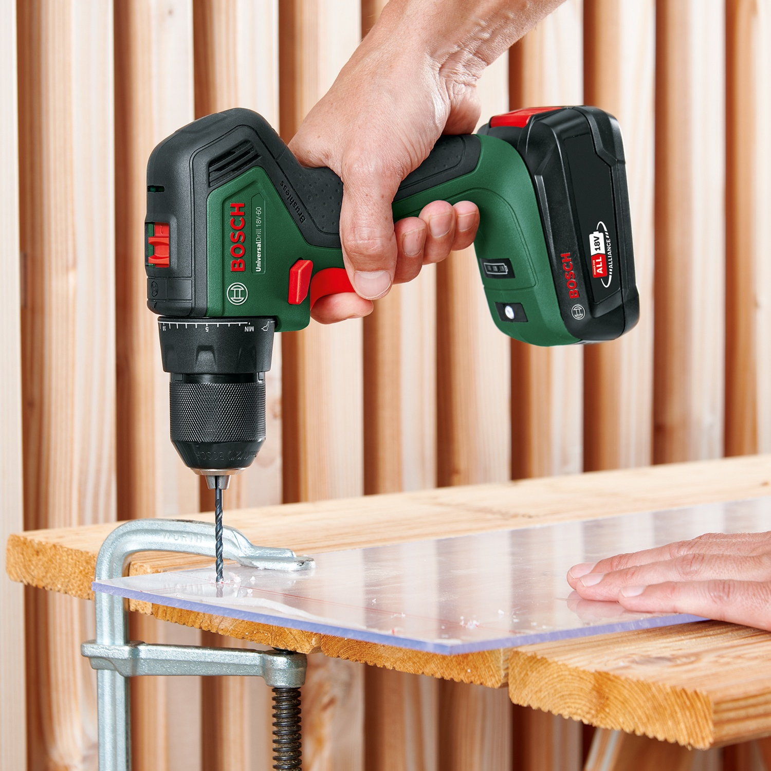 Una persona fora un foro in un foglio di plastica trasparente bloccato a una tavola di legno con un trapano Bosch verde e nero.