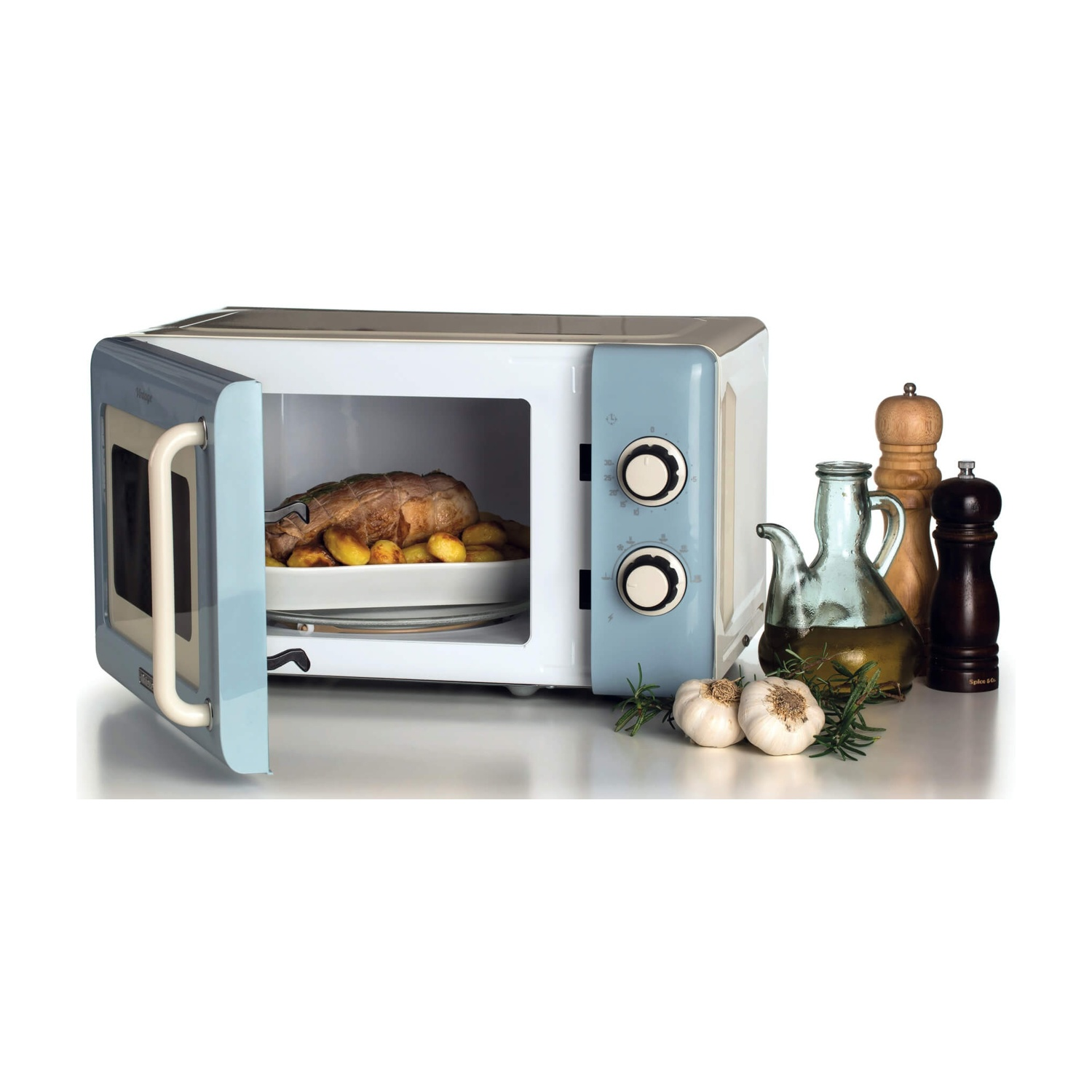 Un forno a microonde retrò con cibo arrosto all'interno. Accanto, olio, spezie ed erbe aromatiche sono posizionati su una superficie bianca.