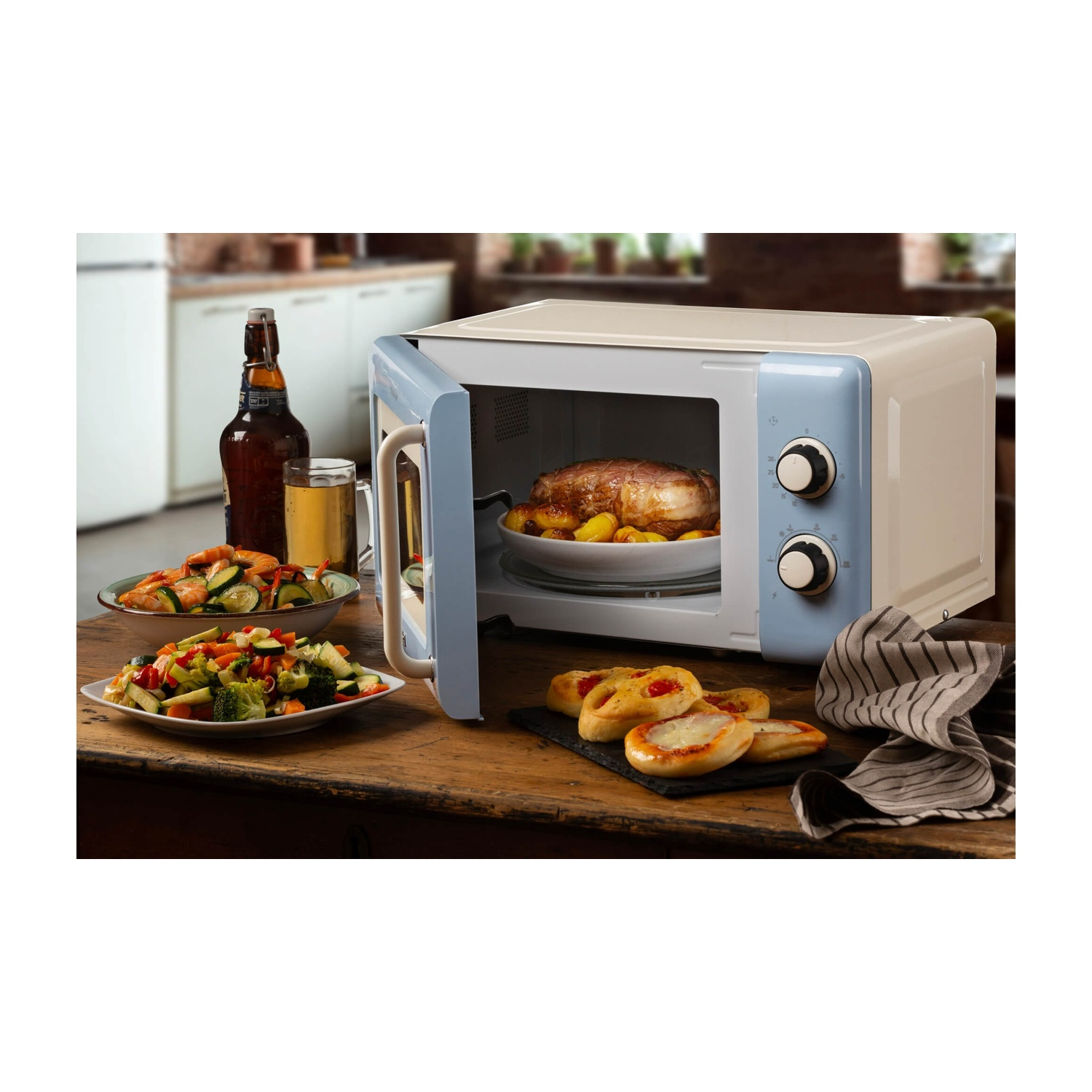 Un microonde con cibo all'interno, vari contorni, birra e mini pizze su un tavolo di legno.