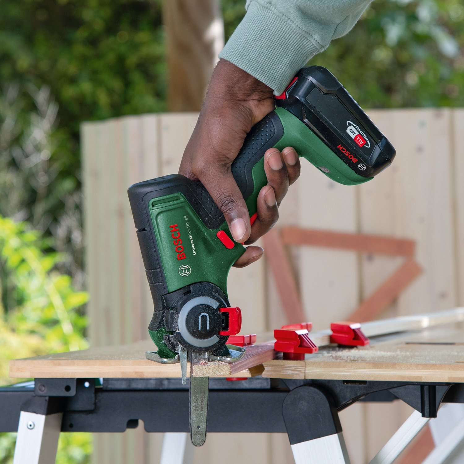 Una persona usa una sega Bosch verde e nera per tagliare un pezzo di legno su un banco da lavoro. Segatura visibile.