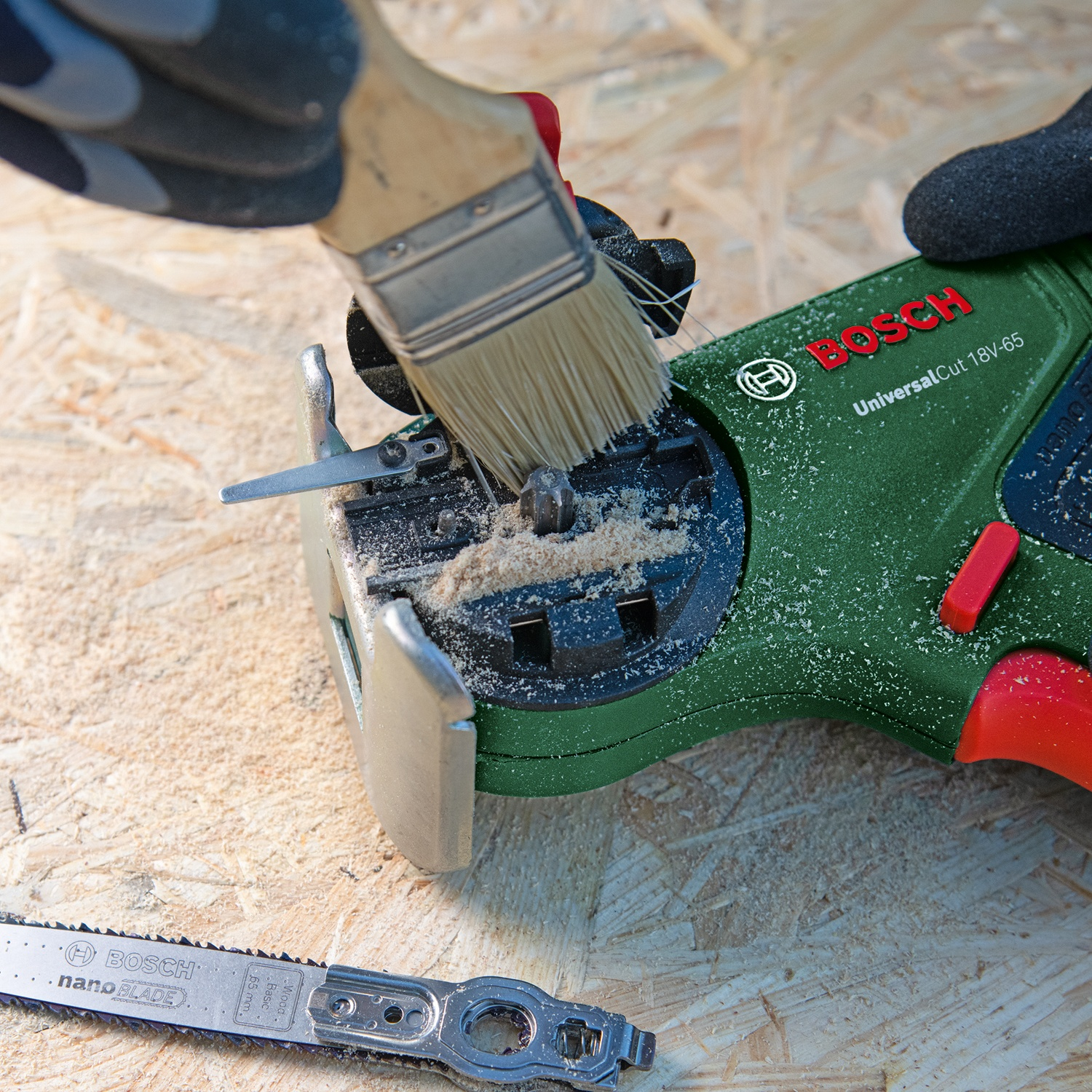Una mano usa un pennello per pulire la segatura da un seghetto alternativo Bosch verde. Una lama e il seghetto sono su una superficie di legno.