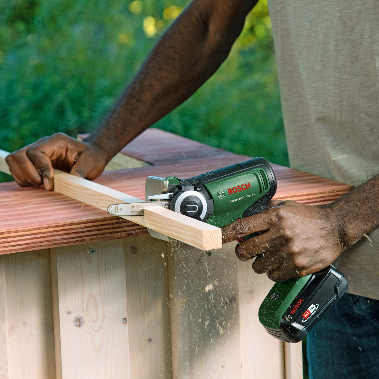 Una persona usa una sega Bosch verde e nera per tagliare una tavola di legno. Segatura visibile. Lo sfondo è verde sfocato.