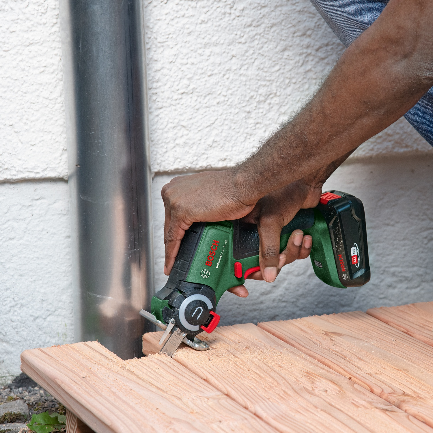 Una persona usa una sega elettrica verde e nera su legno. Sullo sfondo un tubo di metallo e un muro bianco.