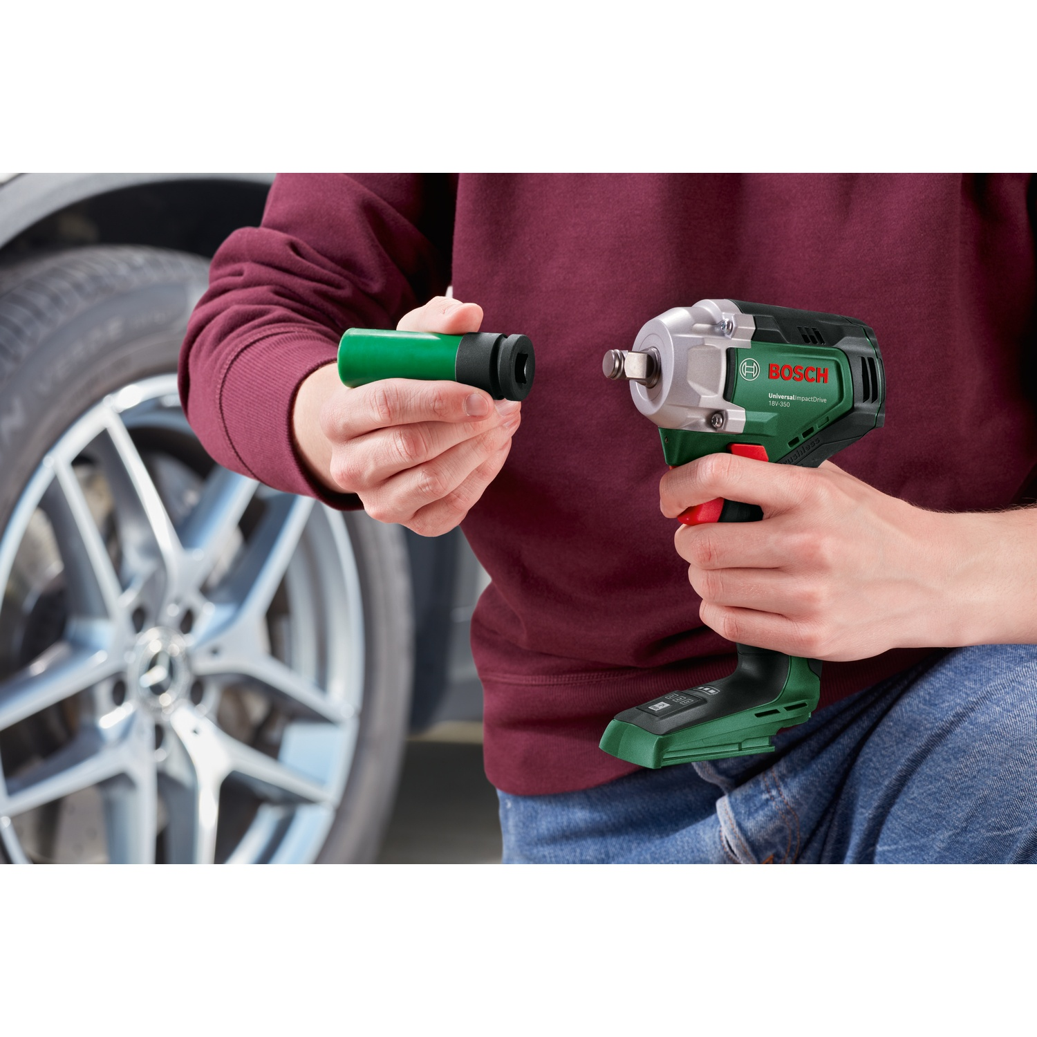 Una persona tiene una chiave a bussola verde e un avvitatore a impulsi Bosch. Sullo sfondo ci sono uno pneumatico e un cerchione dell'auto. La persona indossa una camicia marrone.