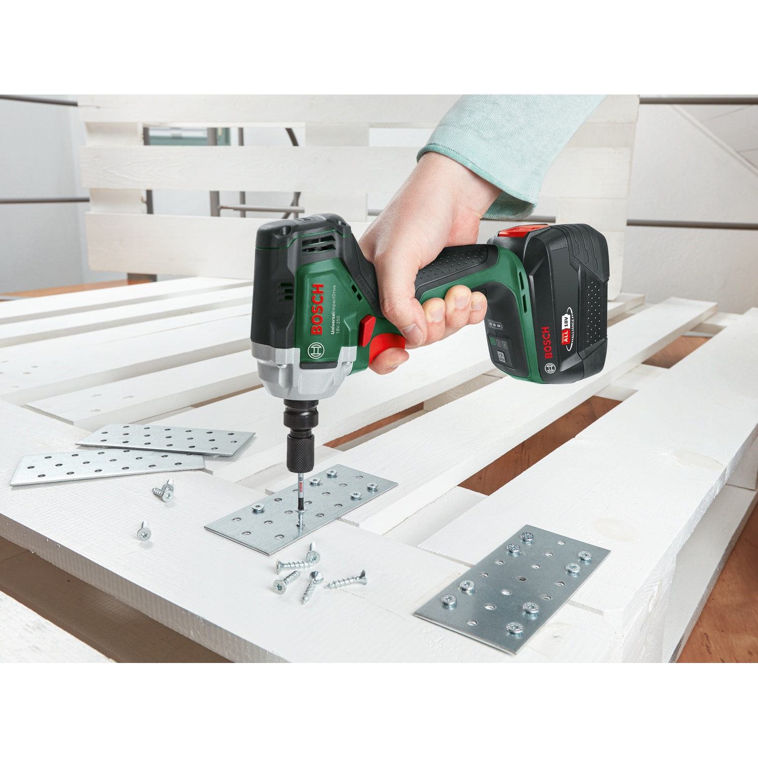 Una mano usa un avvitatore a impulsi Bosch verde e nero per avvitare piastre metalliche a un pallet di legno bianco. Viti e piastre visibili.