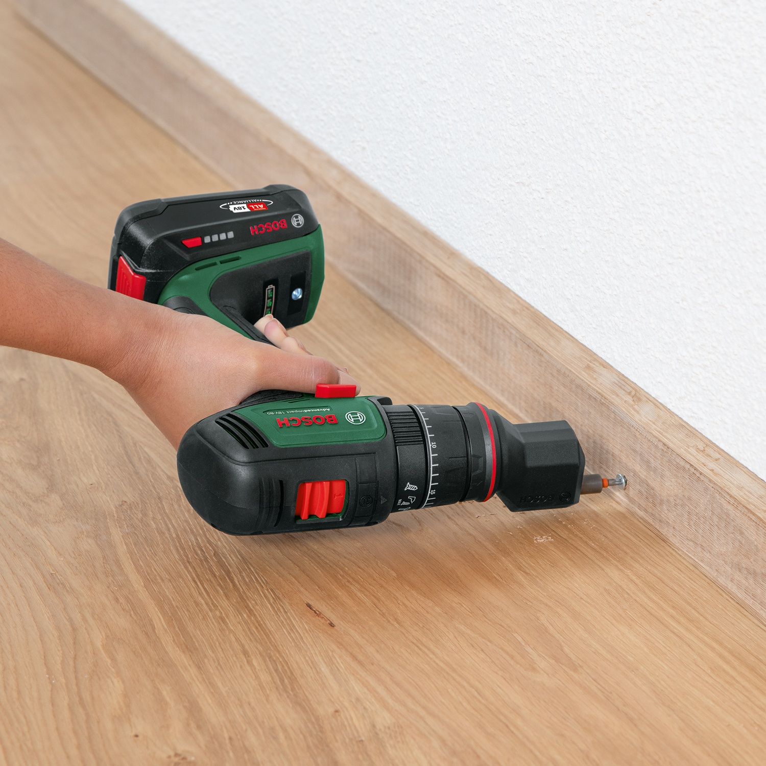 Una persona usa un trapano Bosch per avvitare in un battiscopa di legno. Il trapano è nero e verde, il pavimento è in rovere e il muro è bianco.