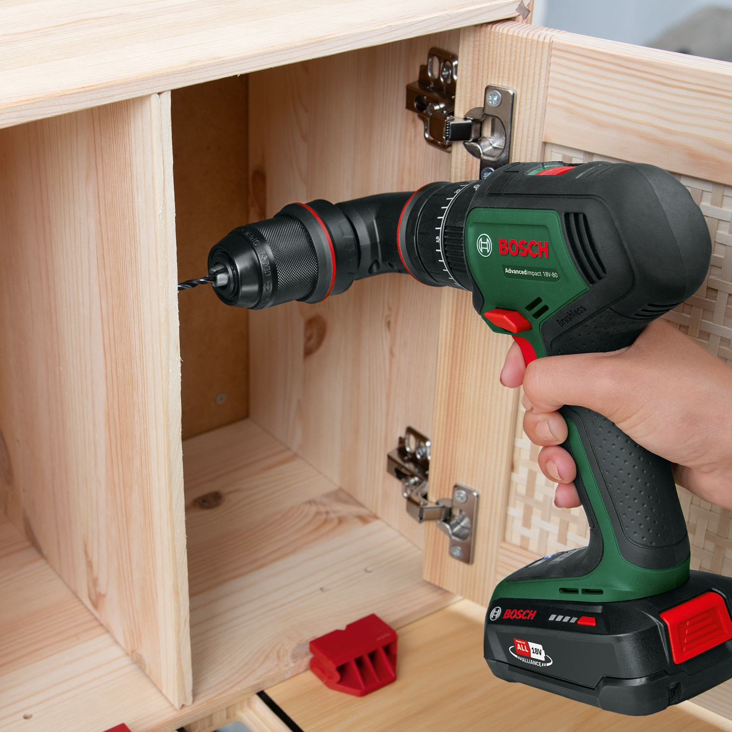 Una mano tiene un trapano Bosch, che fora un mobile di legno. Il trapano è verde e nero. Sono visibili cerniere e clip rosse.