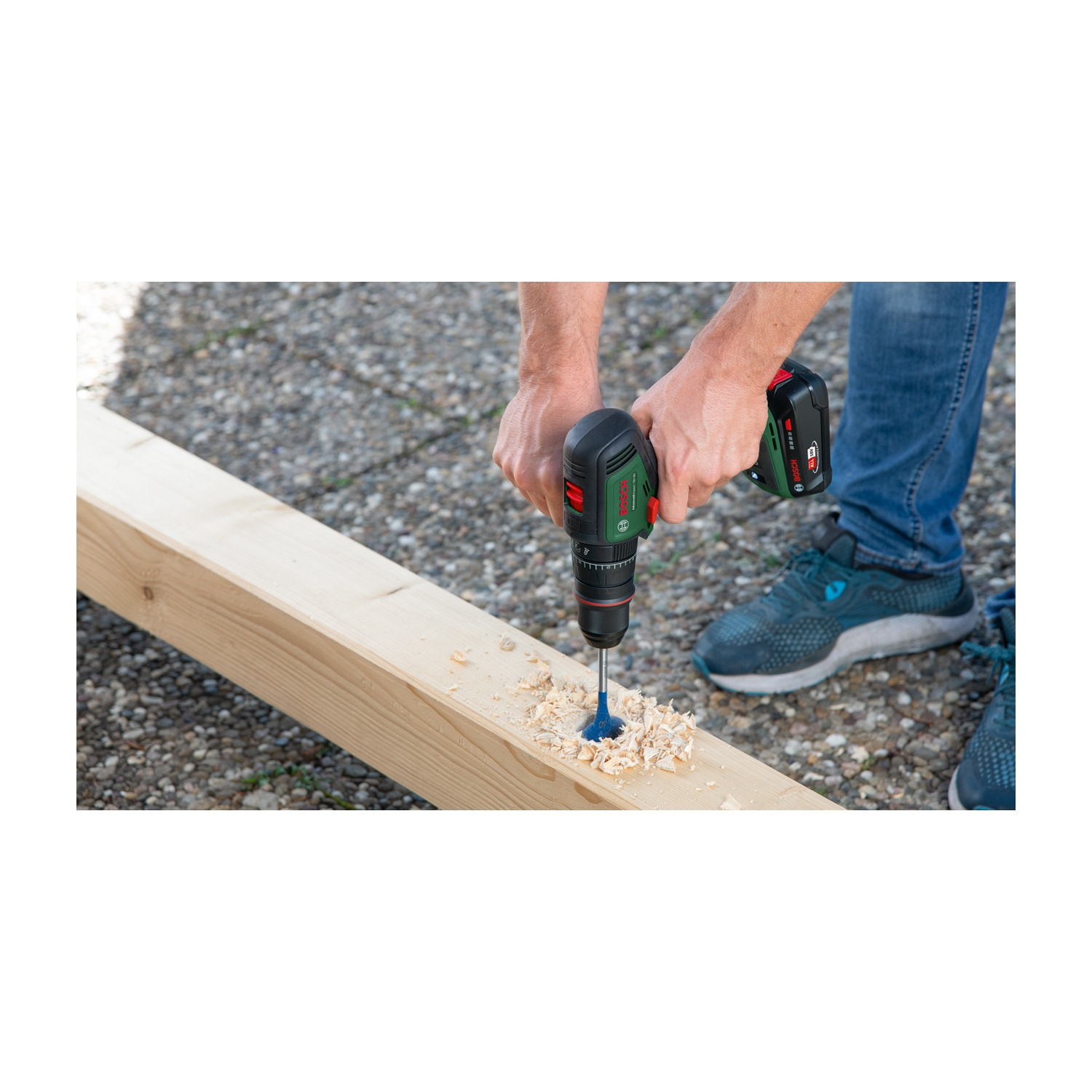 Una persona fora un'asse di legno con un trapano elettrico. Sono presenti trucioli di legno. La persona indossa jeans e scarpe da ginnastica blu.