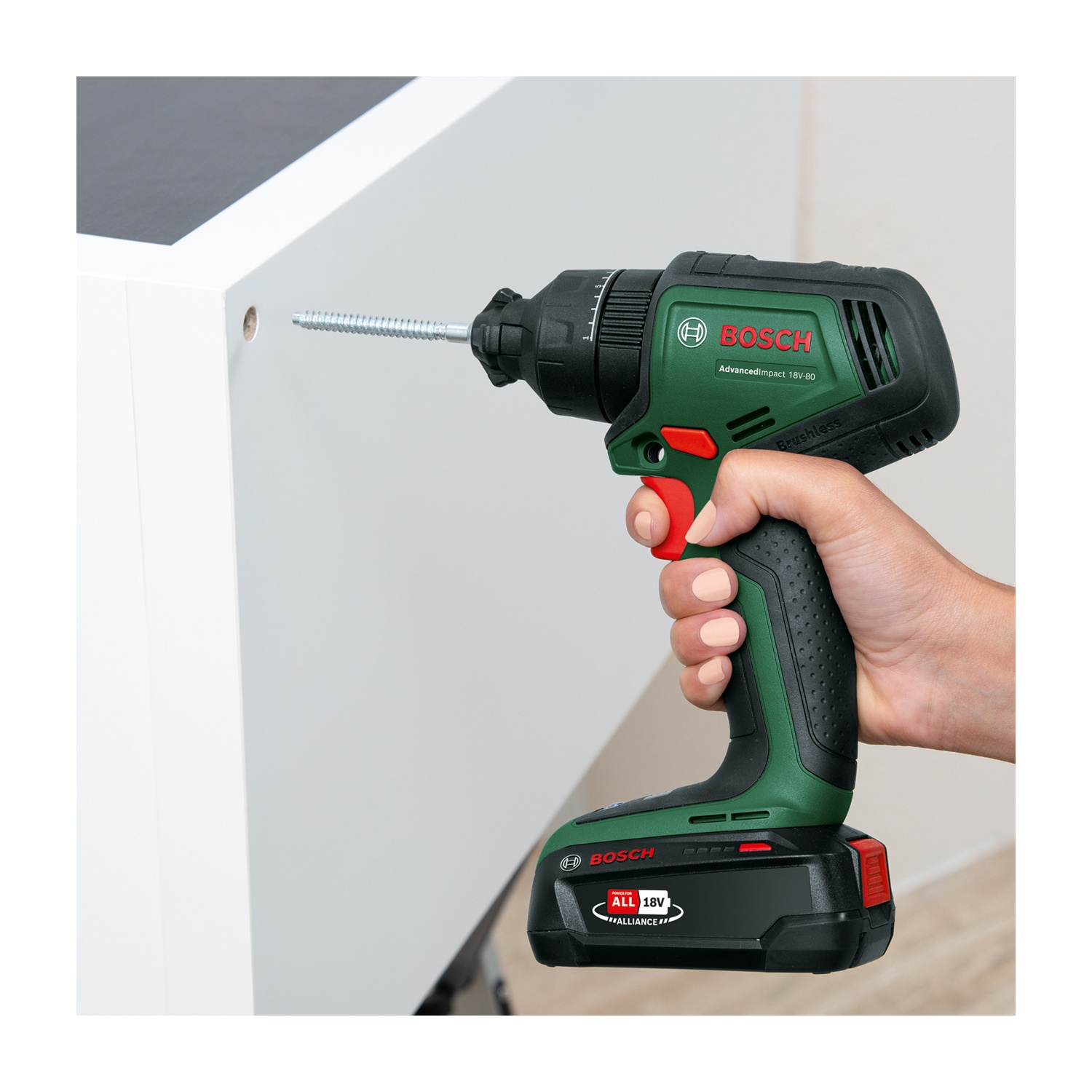 Una mano tiene un trapano Bosch verde e nero, avvitando una vite argentata in un armadio bianco. Lo sfondo è una parete chiara.