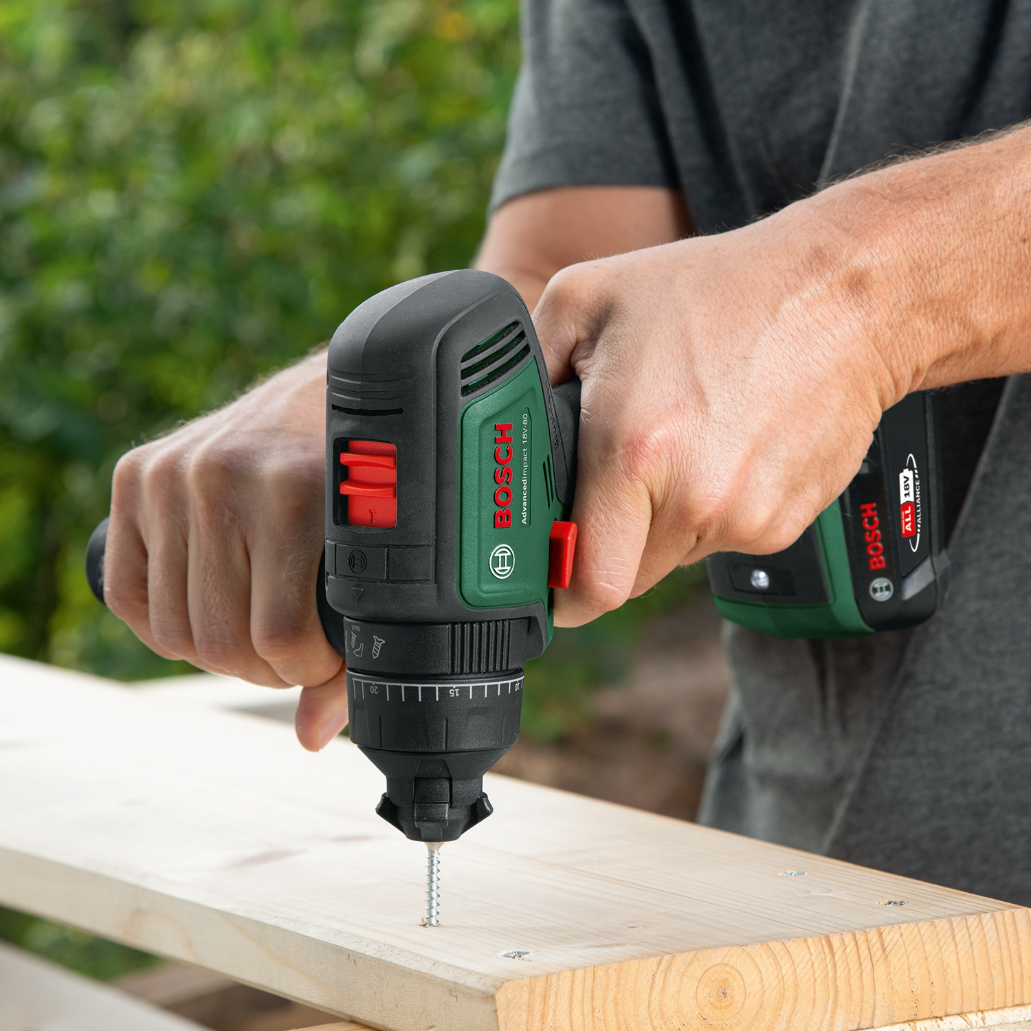 Una persona usa un trapano avvitatore Bosch per avvitare in una tavola di legno. Il trapano è nero e verde. Lo sfondo è verde.