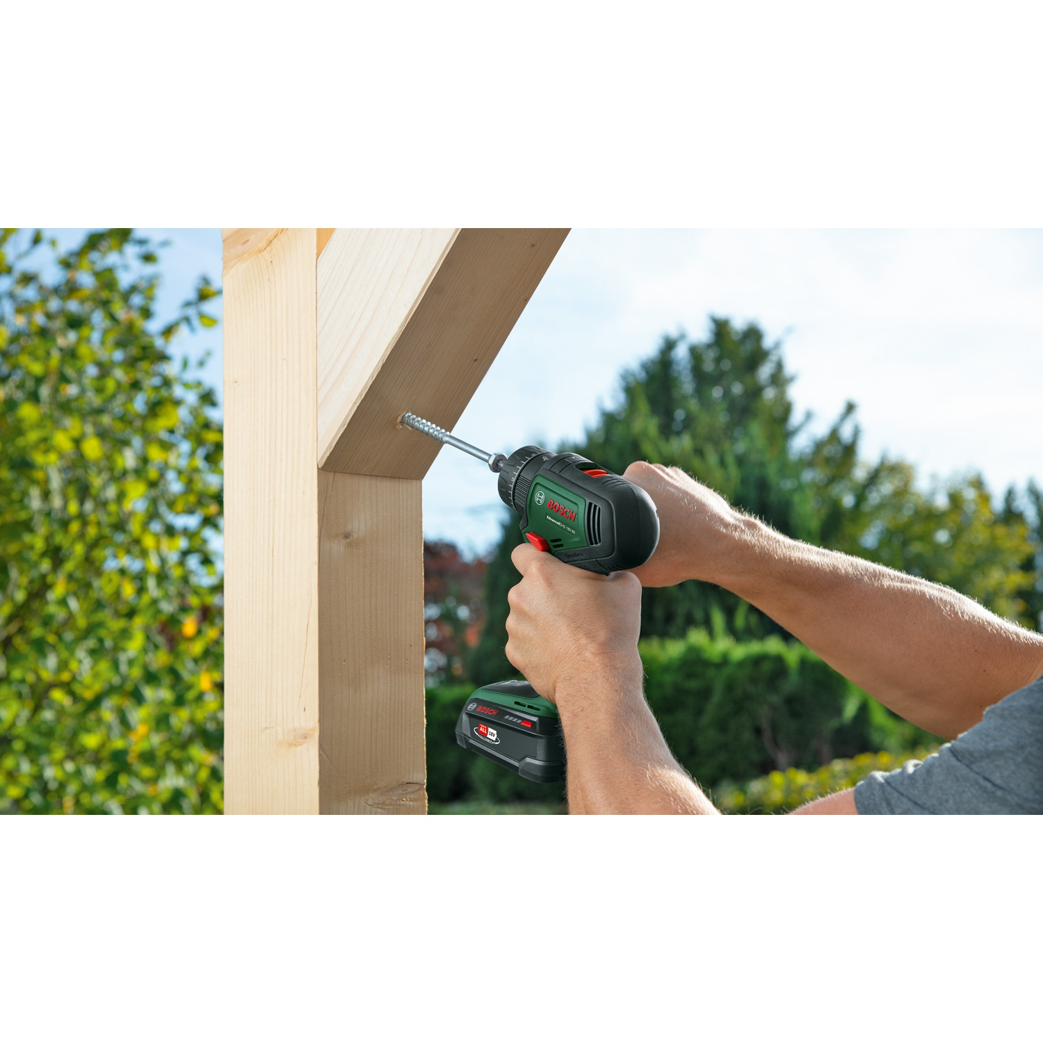 Una persona avvita una vite nel legno con un trapano avvitatore verde e nero. Lo sfondo include alberi e cielo.