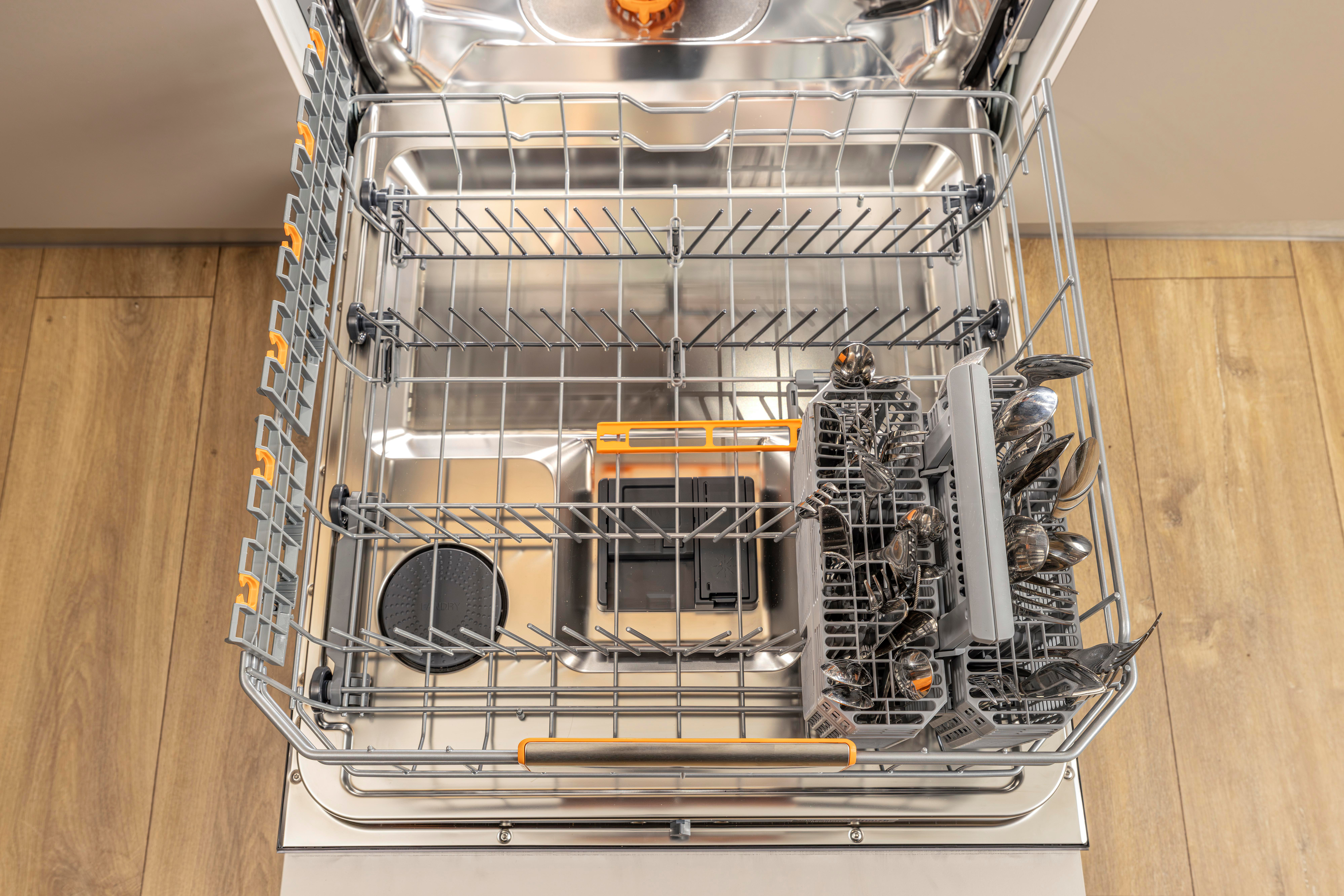 Ein offener Geschirrspüler mit Besteck im Korb. Der Geschirrspüler ist aus Edelstahl mit orangefarbenen Akzenten. Holzboden im Hintergrund.
