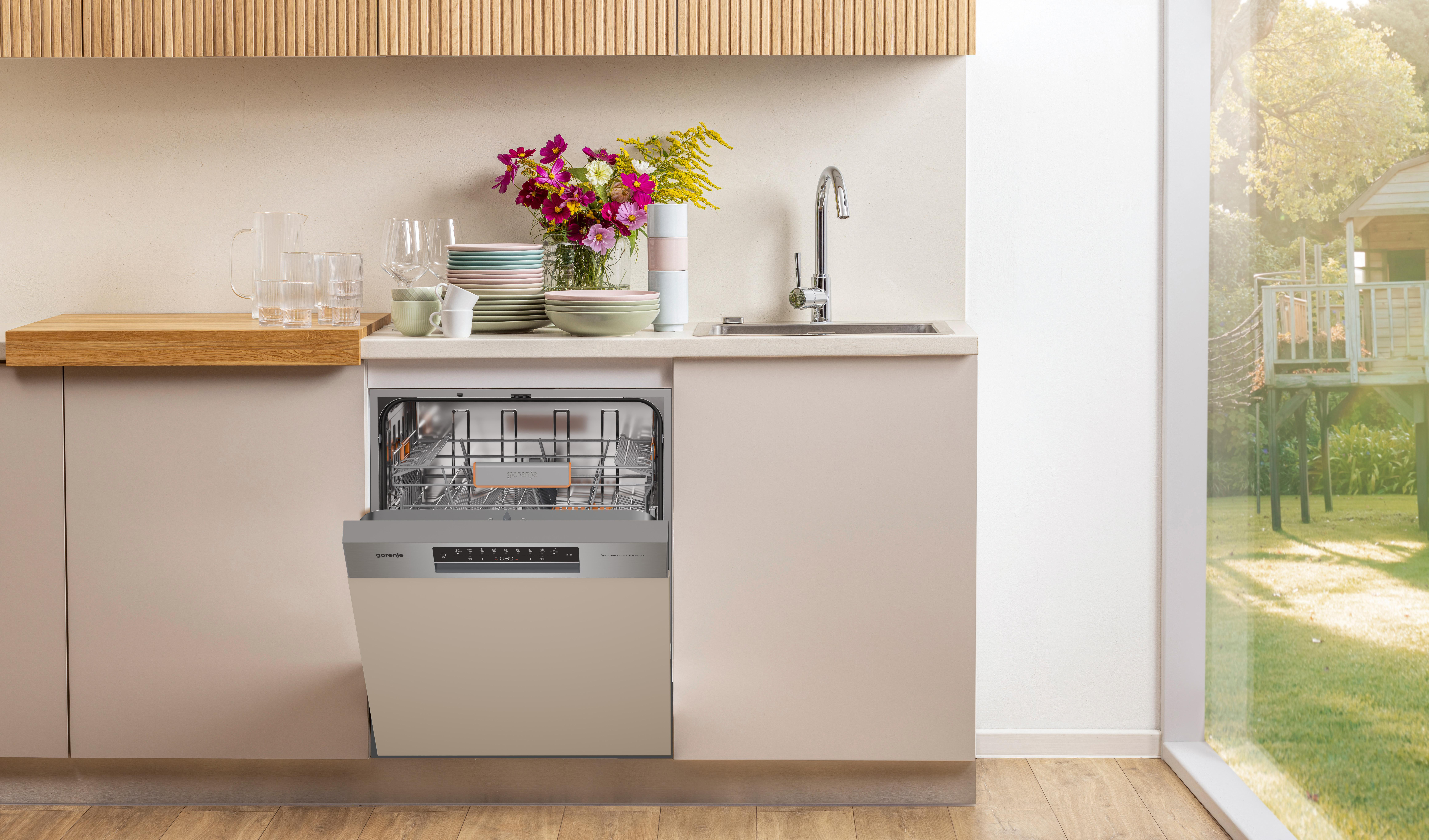 Küche mit geöffneter Spülmaschine, Blumen, Geschirr und Spüle. Fenster zeigt Baumhaus draußen.