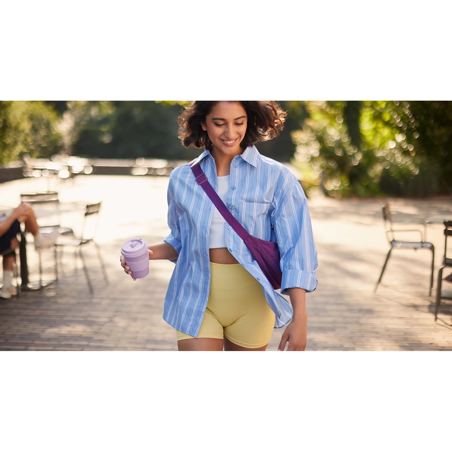 Una donna sorride in un parco, tenendo una tazza viola. Indossa una camicia blu a righe, pantaloncini gialli e una borsa viola.