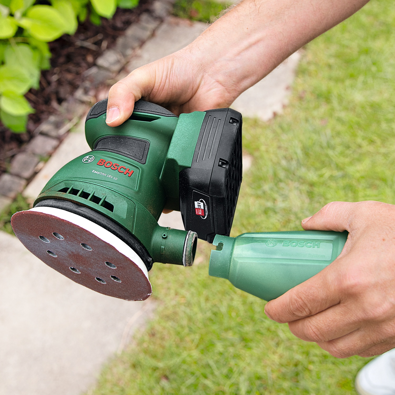 Persona che tiene in mano una levigatrice Bosch verde e un raccoglitore di polvere. Il tampone di levigatura è marrone. Lo sfondo è erba e fogliame.