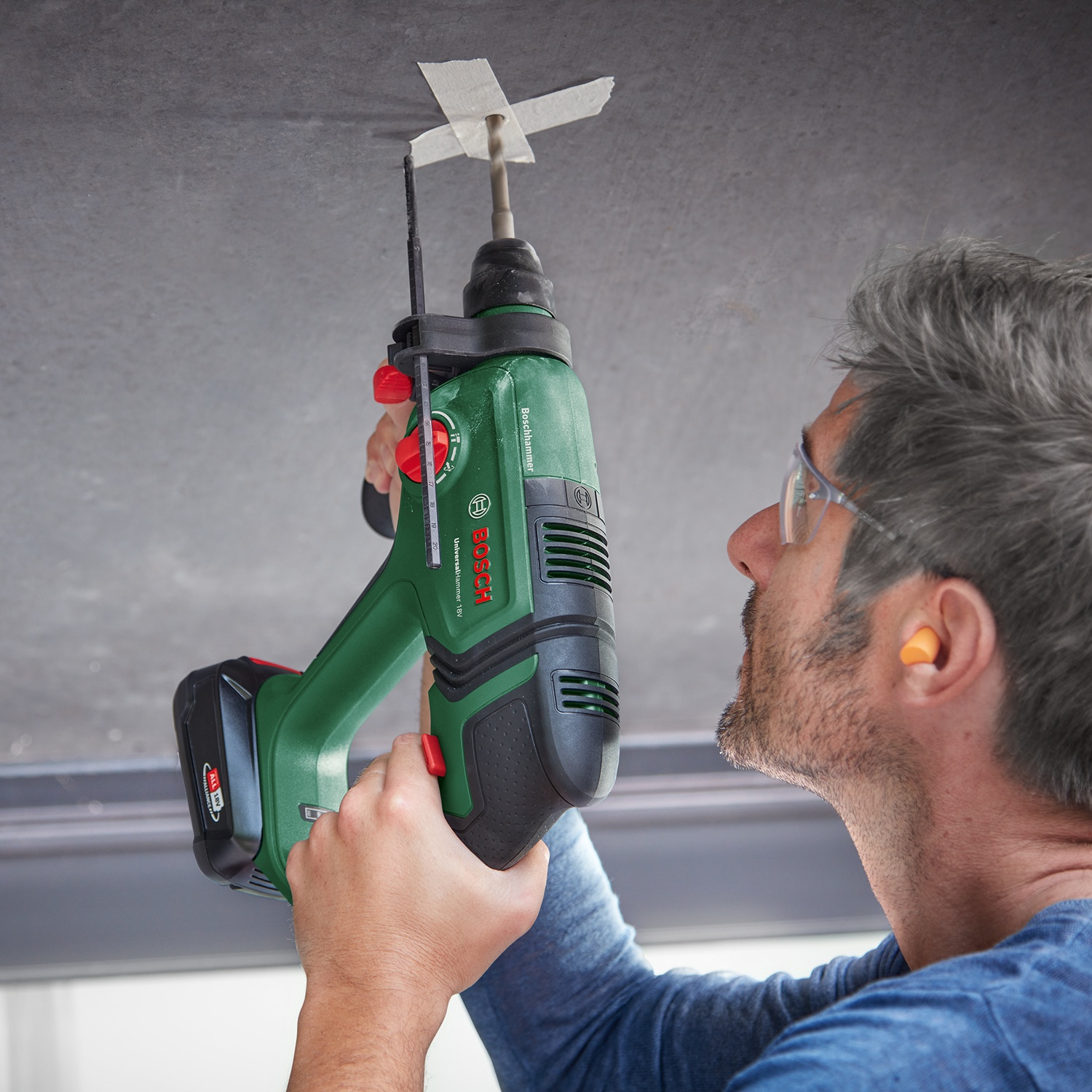 Un uomo fora un soffitto con un martello perforatore Bosch verde. Indossa occhiali di sicurezza e tappi per le orecchie. Nastro adesivo segna il punto.